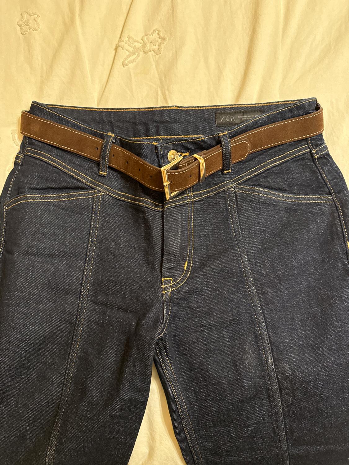 Dark blue denim jeans with visible stitching and a brown belt on a light surface with floral embroidery.