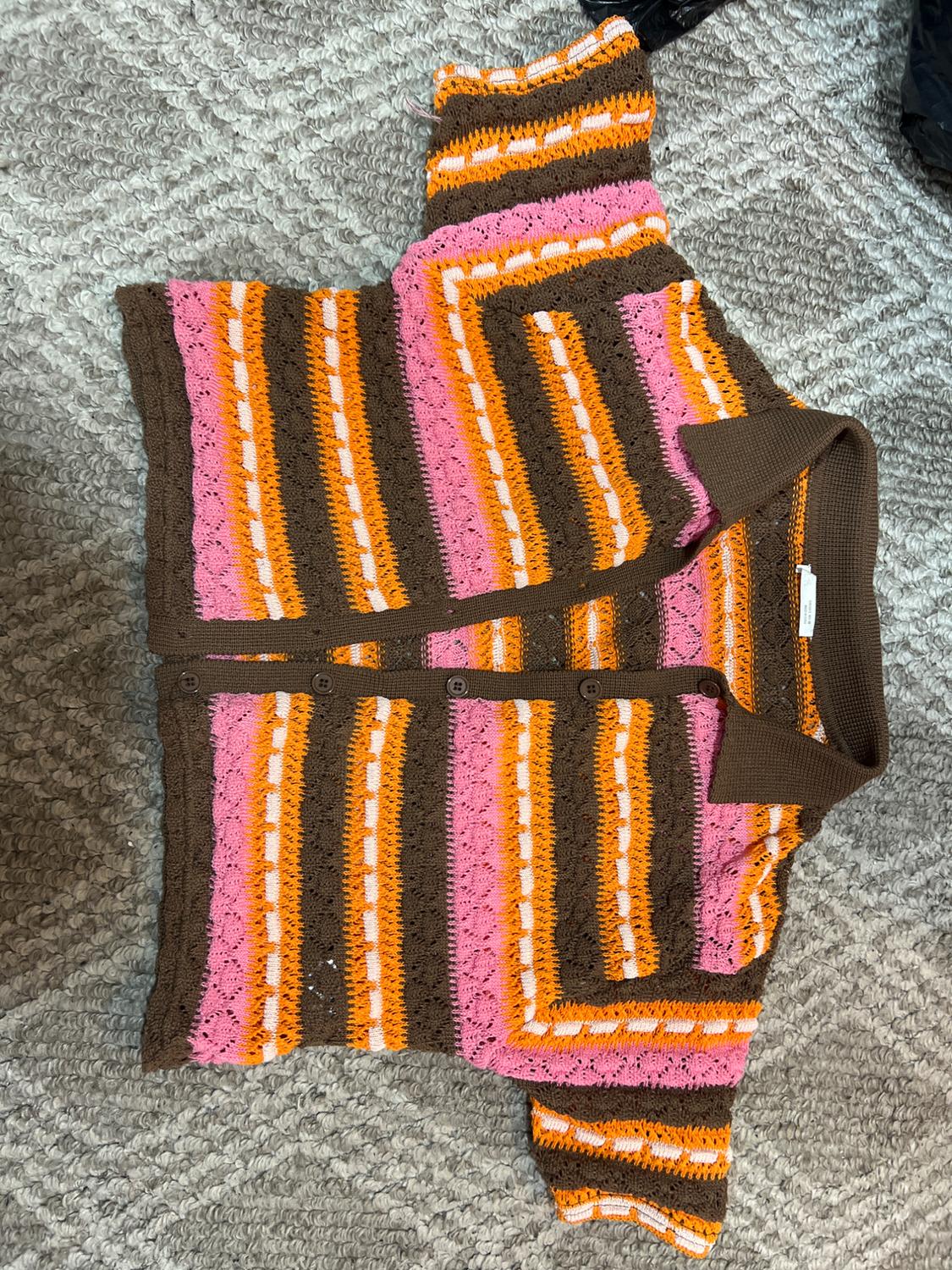 Crochet cardigan with horizontal stripes in pink, orange, brown, and white, on a grey rug with a geometric design.