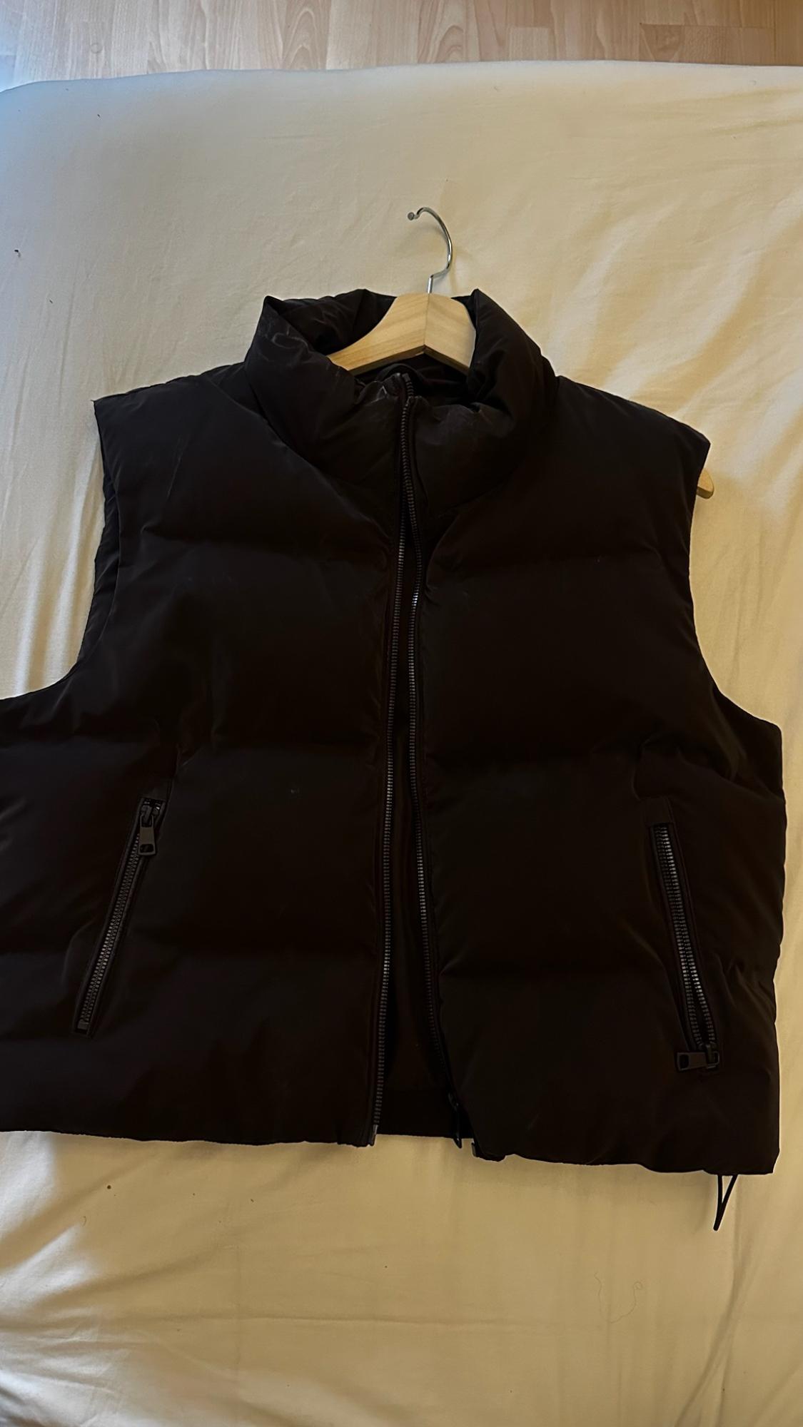 Black padded gilet with a high collar, zip fastening and side pockets with zips. Displayed on a wooden hanger against a light-coloured fabric.