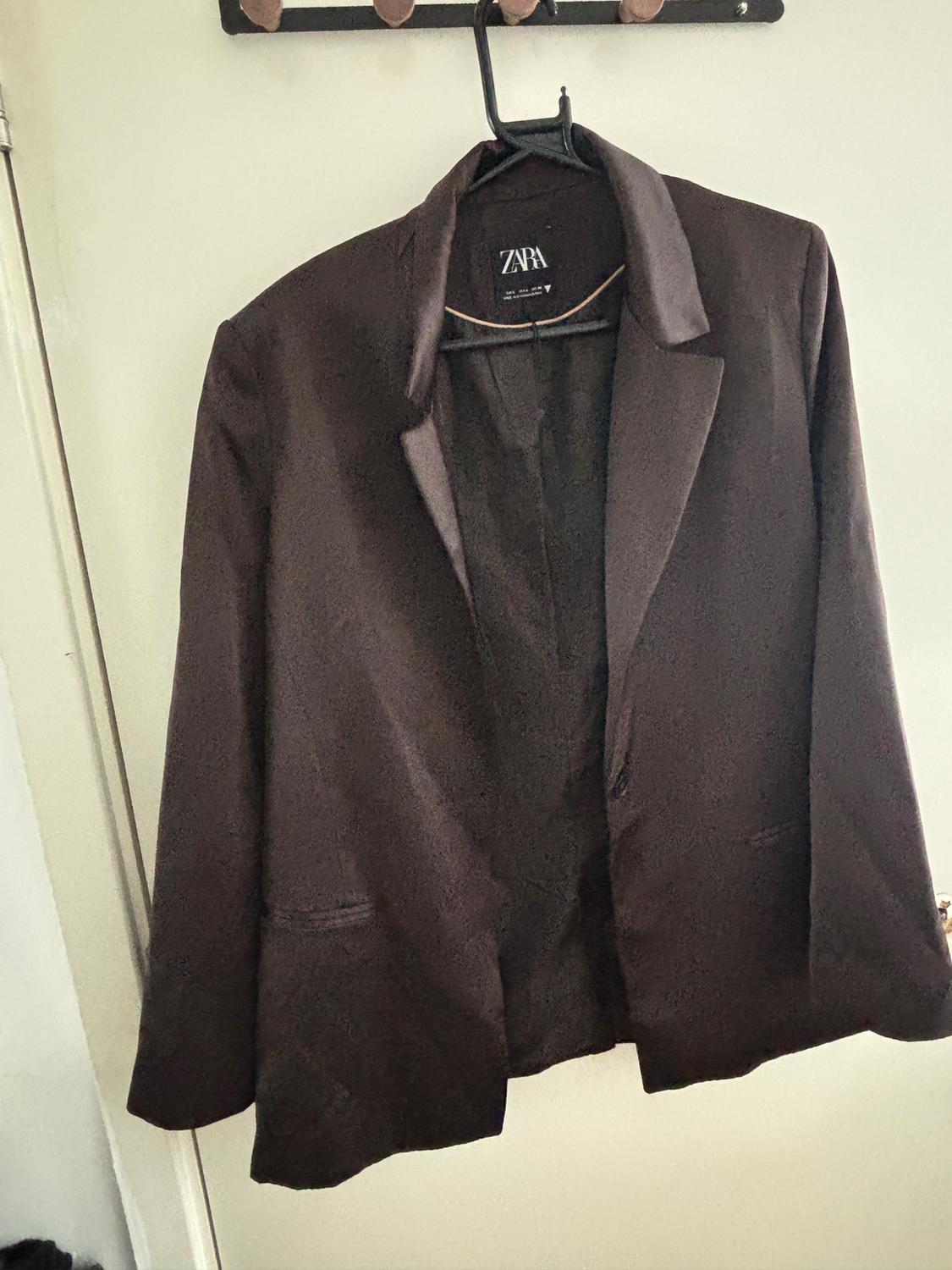 Black jacket hanging on a hanger, with a visible label that reads "ZARA". White background and hook on the wall.