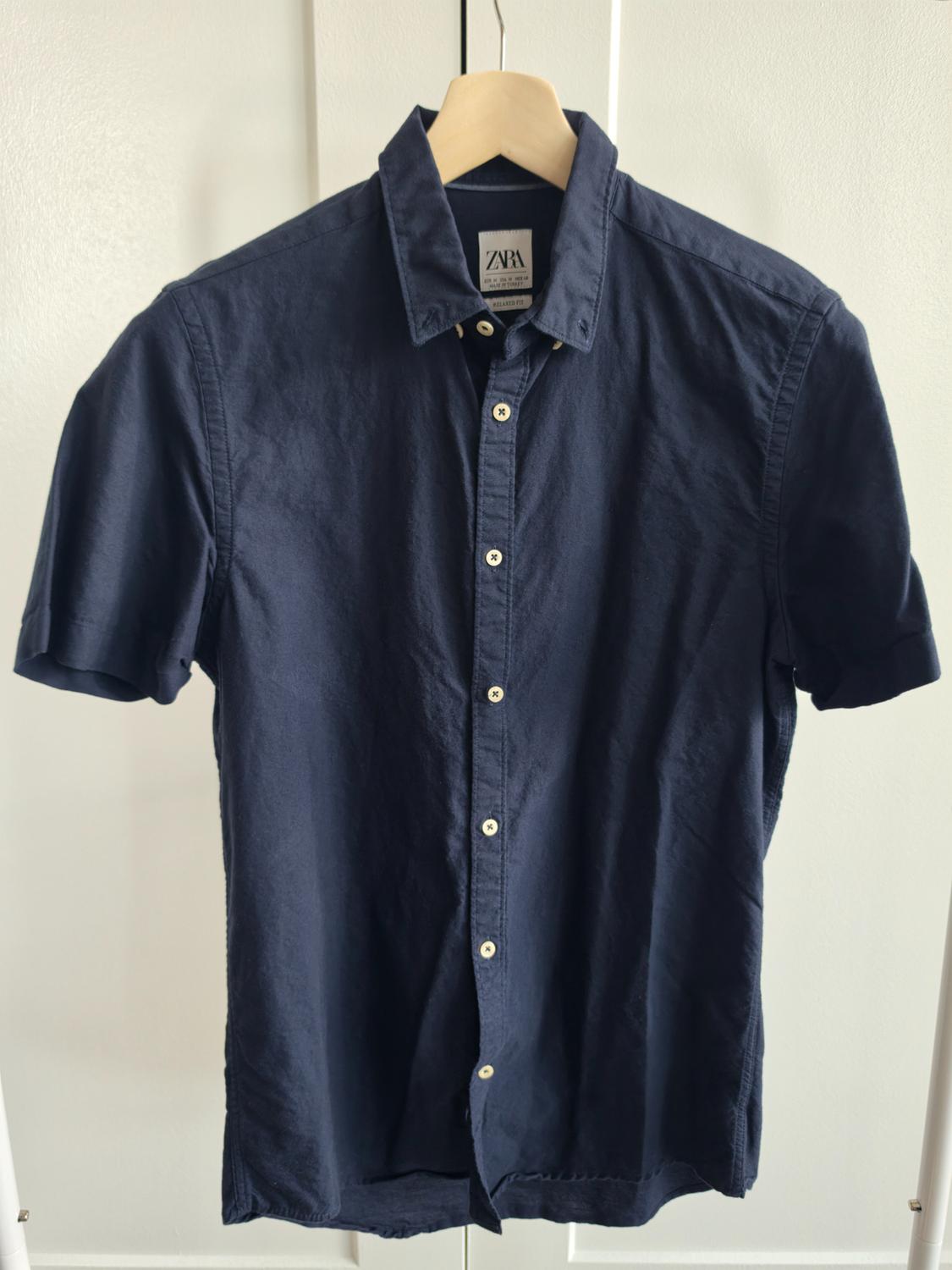 Short-sleeve navy blue button-up shirt, hanging on a wooden hanger against a white background. Label with the text ZARA.