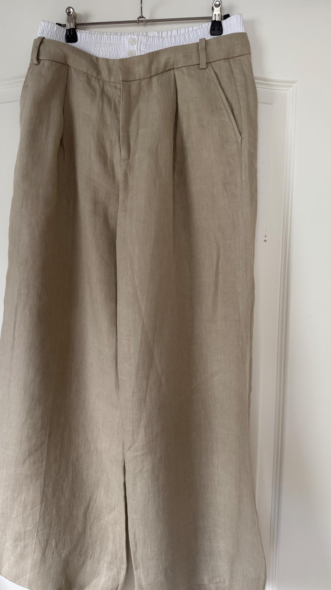 Beige linen trousers hung on a hanger, with a white elasticated waist and visible buttons at the top.