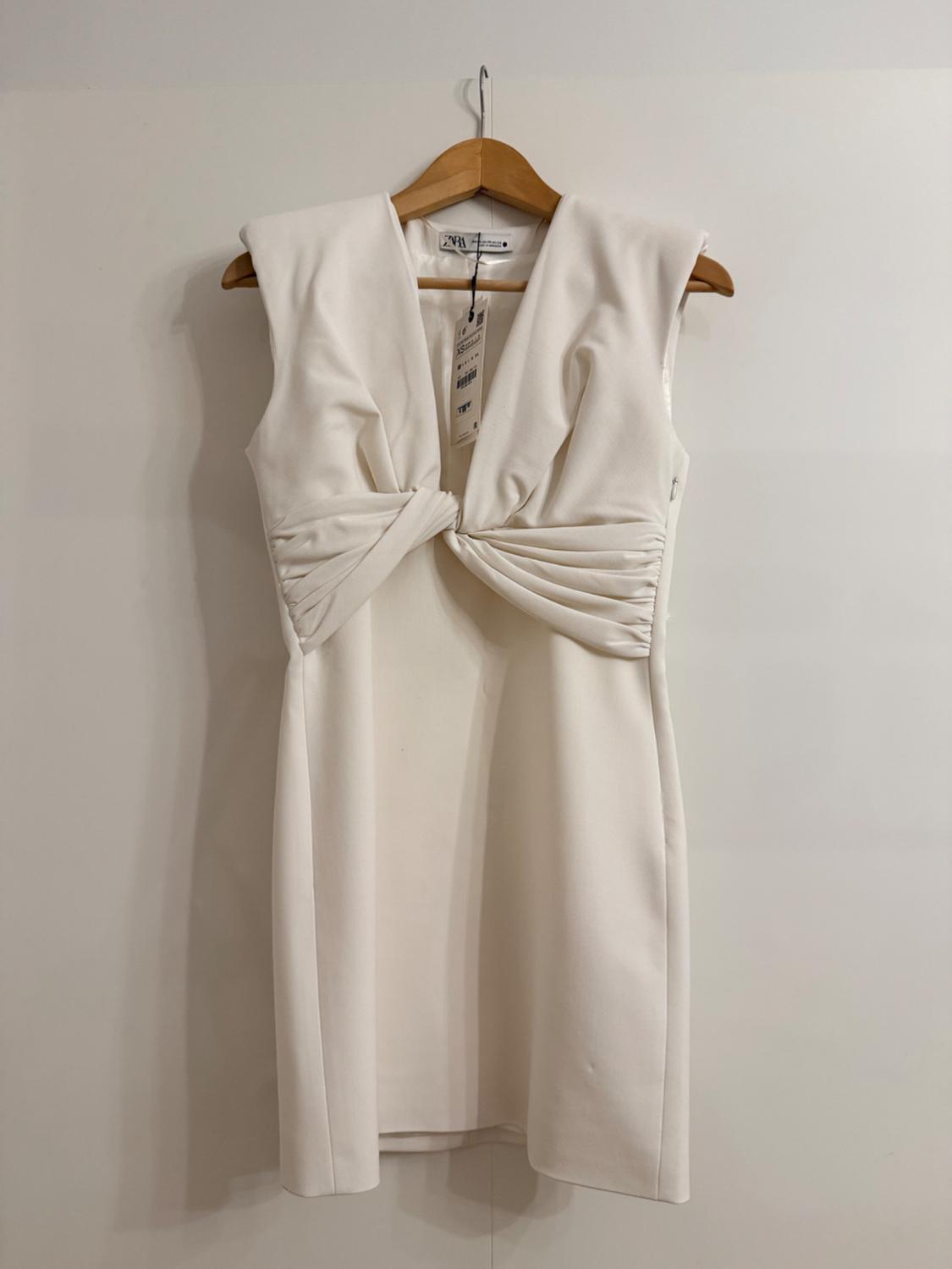 White dress with shoulder pads, a V-neckline and a knot detail at the waist. Hanging on a wooden hanger against a white background.