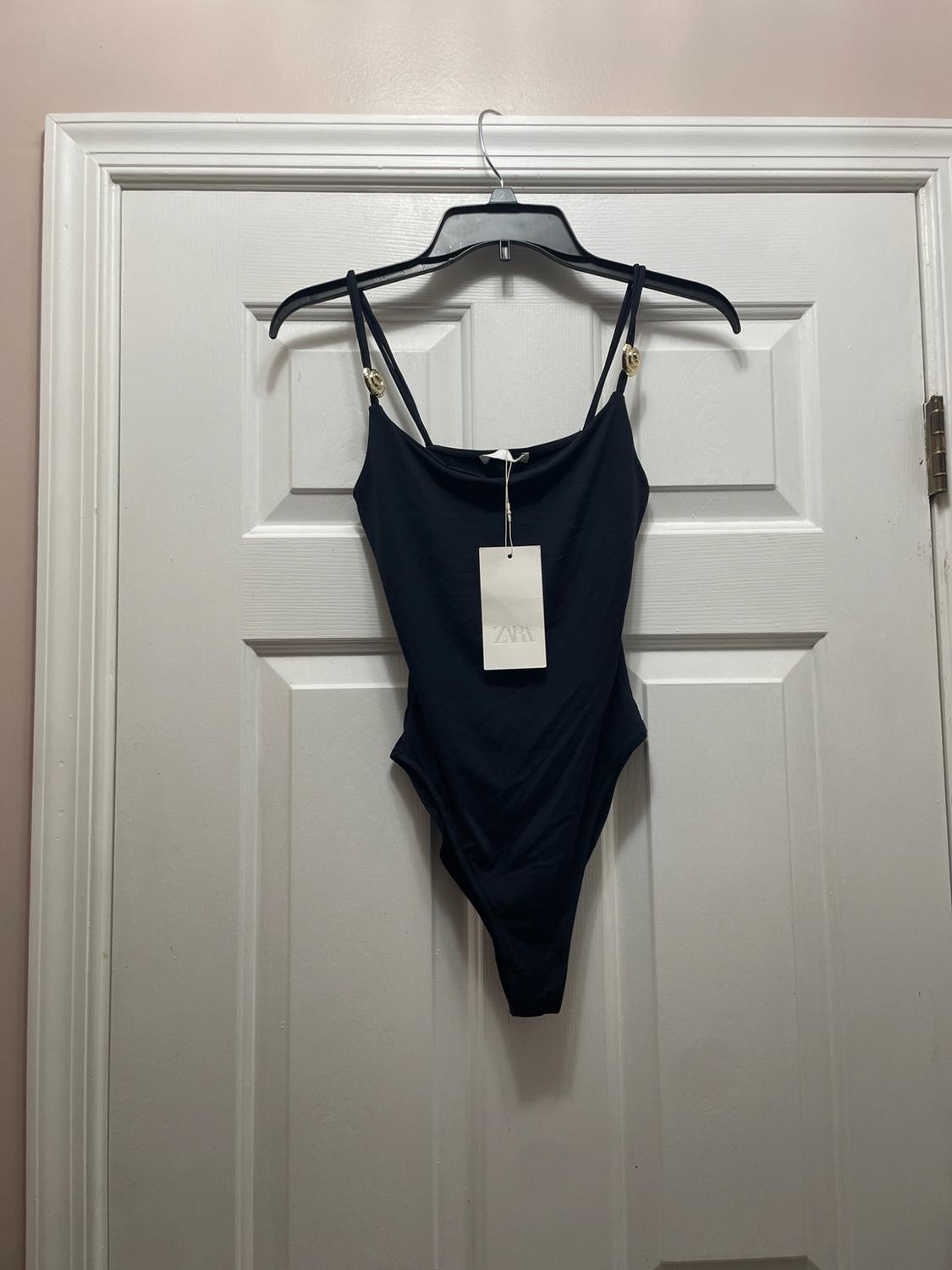 Black swimsuit hanging on a hanger over a white door. It has thin straps with gold rings and a tag with the text ZARA.