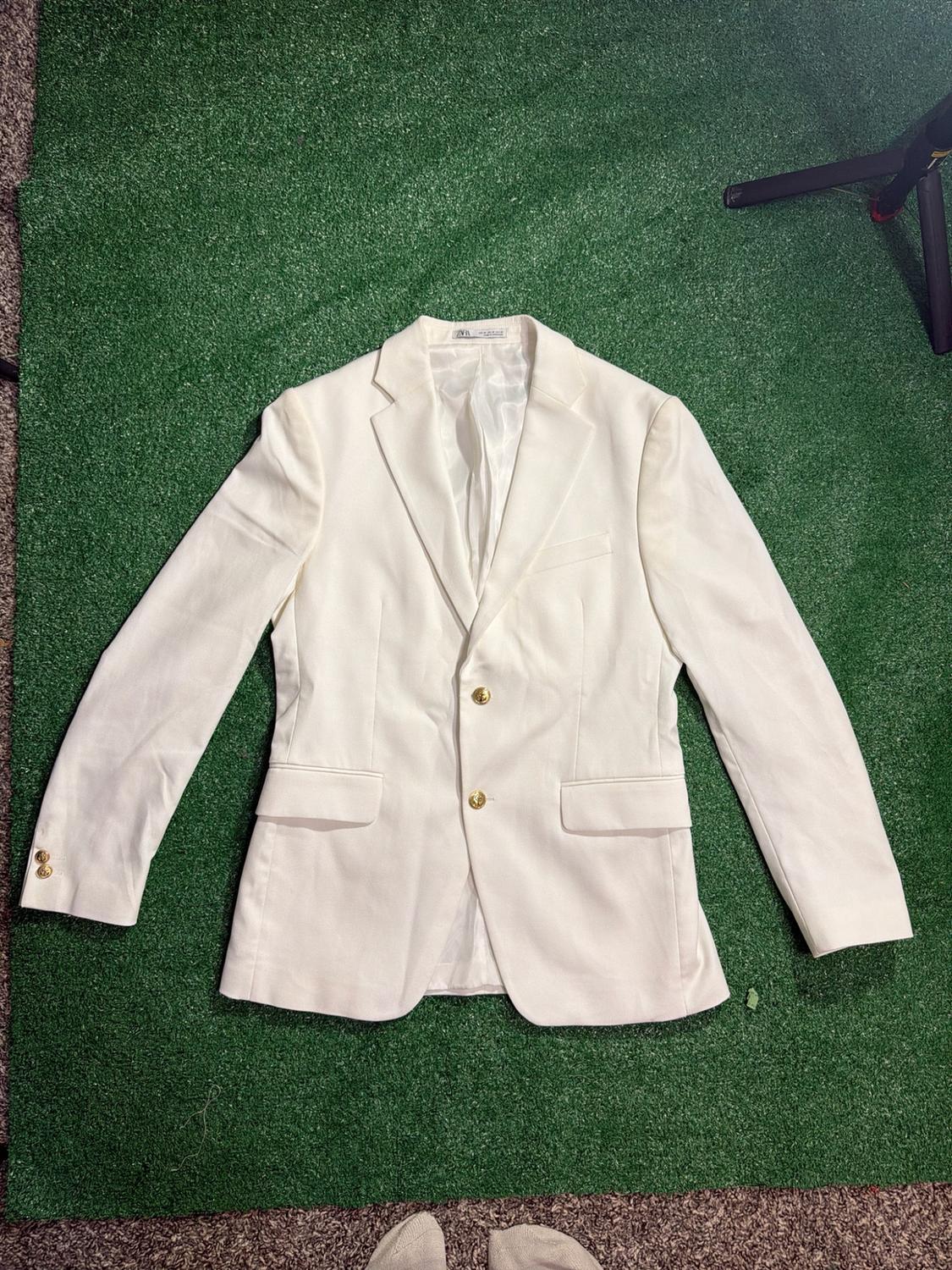 White blazer with gold buttons on green artificial grass. ZARA label. Feet wearing socks and one leg of a tripod are visible.