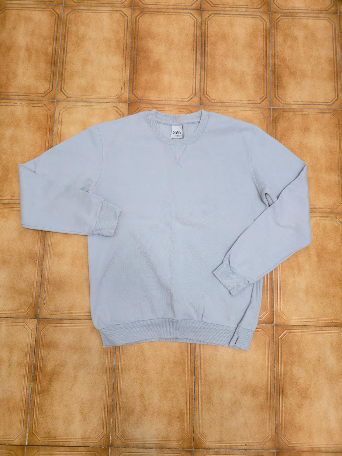 Light blue sweatshirt with a crew neck and long sleeves, placed on a brown tiled floor. Visible label on the collar.