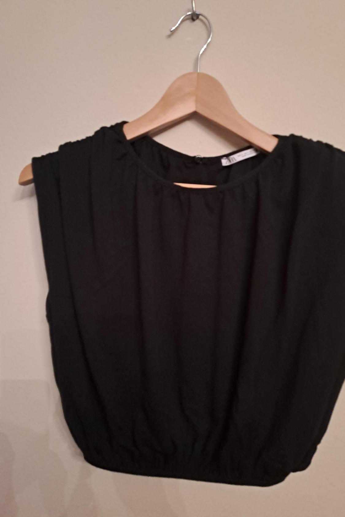 Front view of a black sleeveless blouse with a round neck and ruching on the bottom hem.
