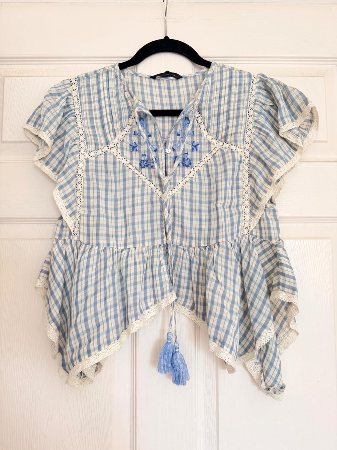 Short-sleeve blouse with a blue-and-white checkered pattern, floral embroidery, lace, an asymmetrical ruffled hem with tassels, against a white door.