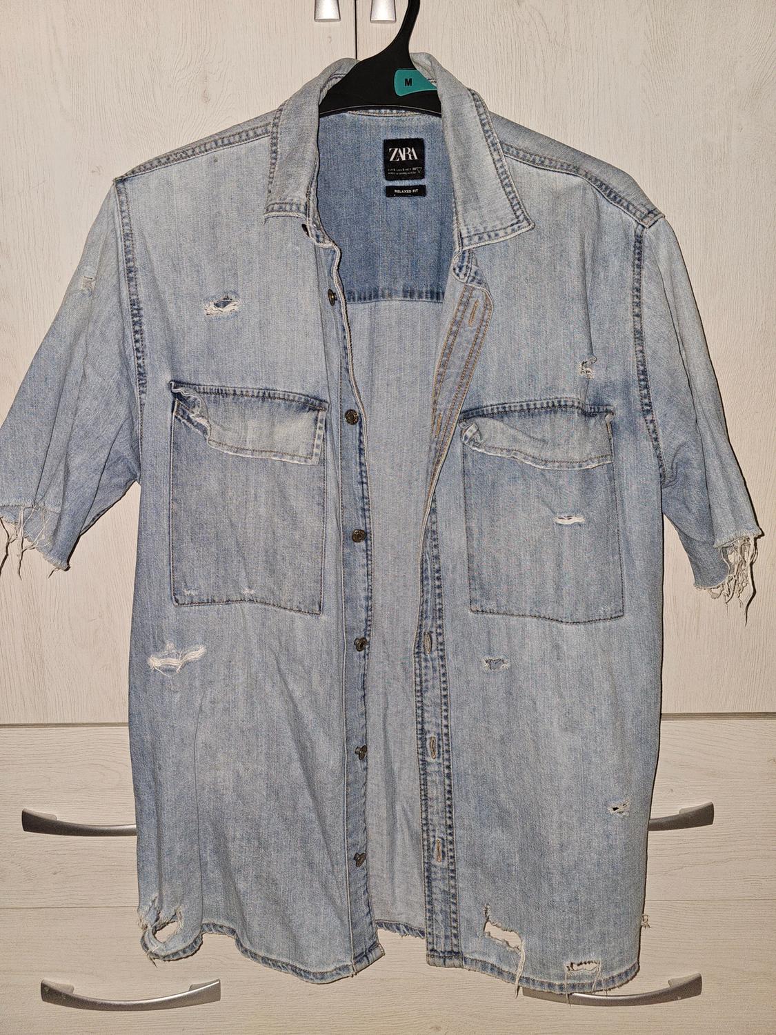 Light blue denim shirt with distressed effect and rips, hanging on a black hanger with "ZARA" label and size "M".