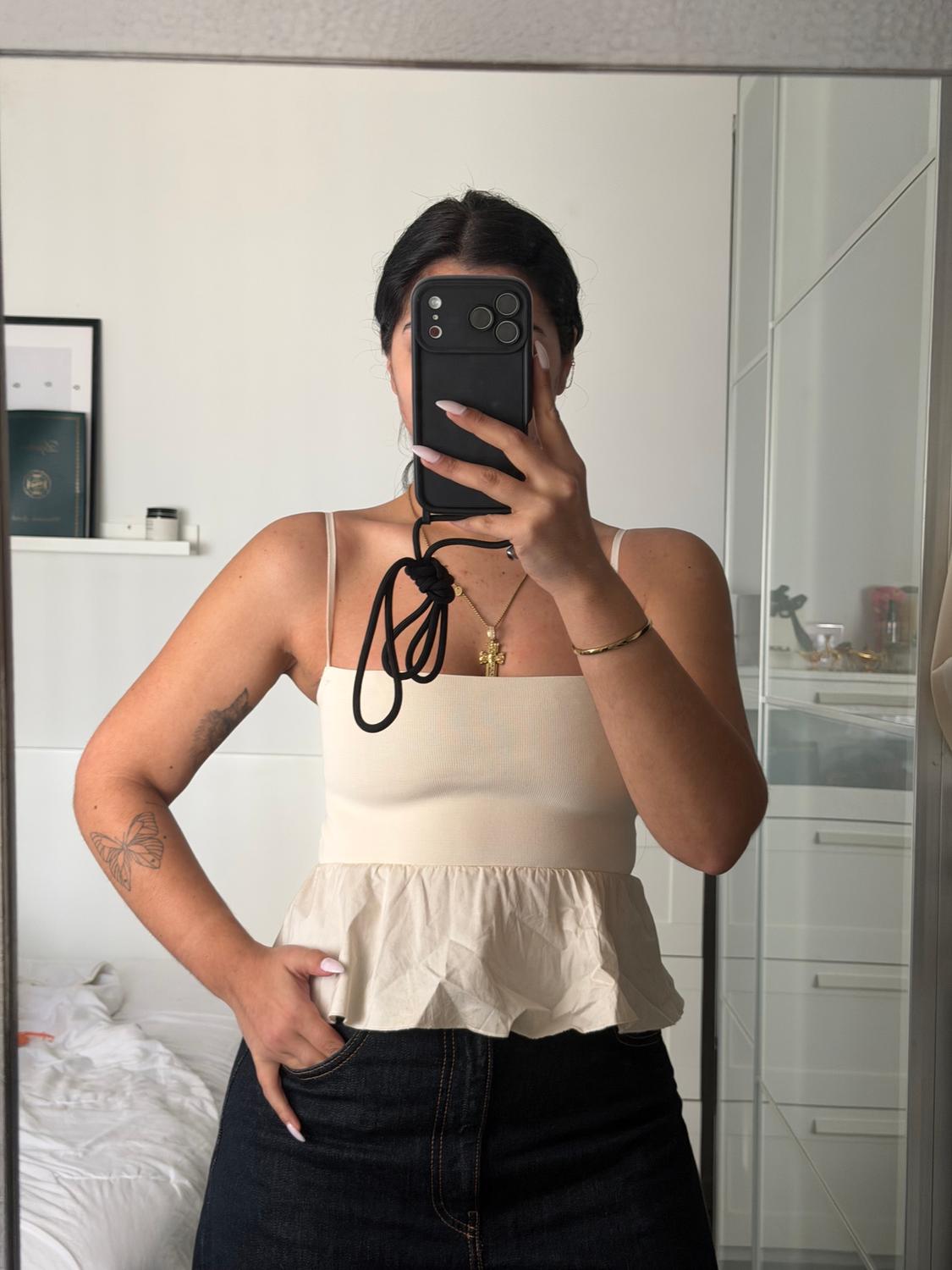 Woman in a beige strappy top and denim skirt, taking a selfie in a mirror. Room with a bed and shelf in the background.