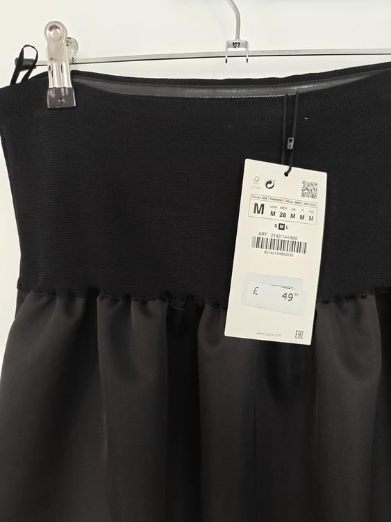 Black skirt hanging on a hanger with a price tag of £49.95 and size M. Smooth, elastic fabric at the top.