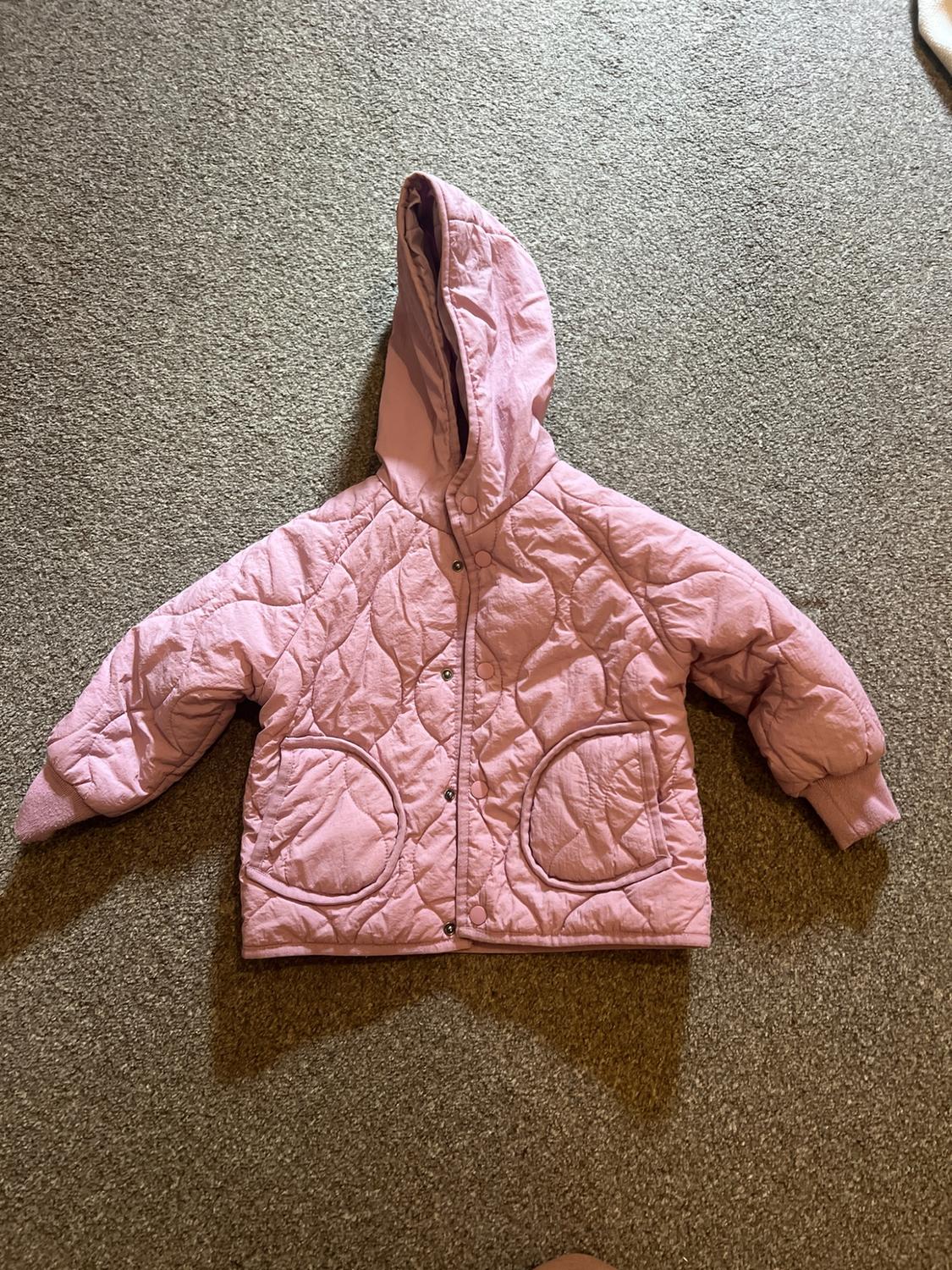 Pink padded hooded jacket with press-stud fastening and front pockets, displayed on a textured rug.
