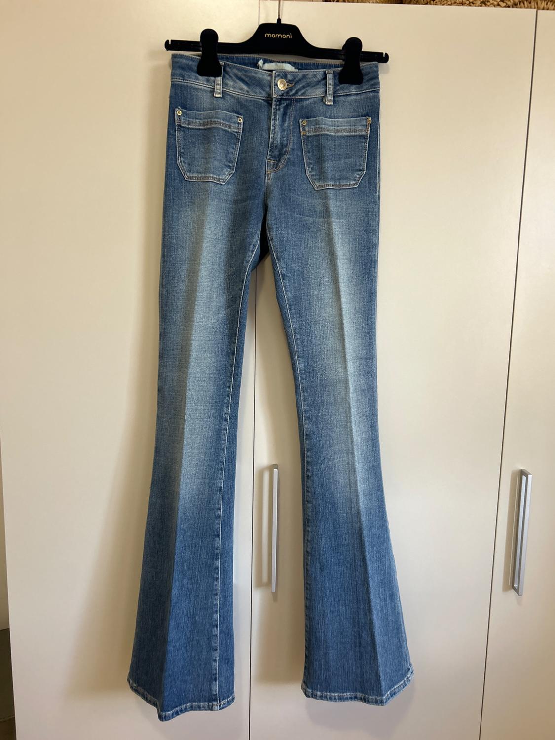 Blue flared denim jeans with front pockets, hanging on a black hanger with the text 'momoni' in front of a white wardrobe.