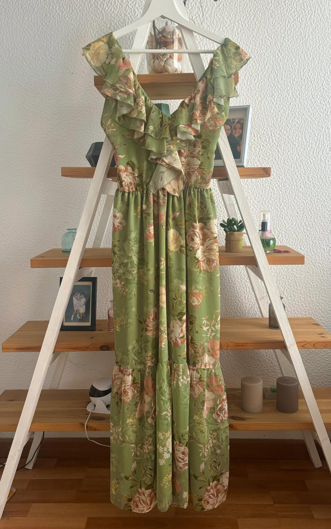 Green long dress with floral print and ruffles on the shoulders, hanging on a wooden shelf with decorations.