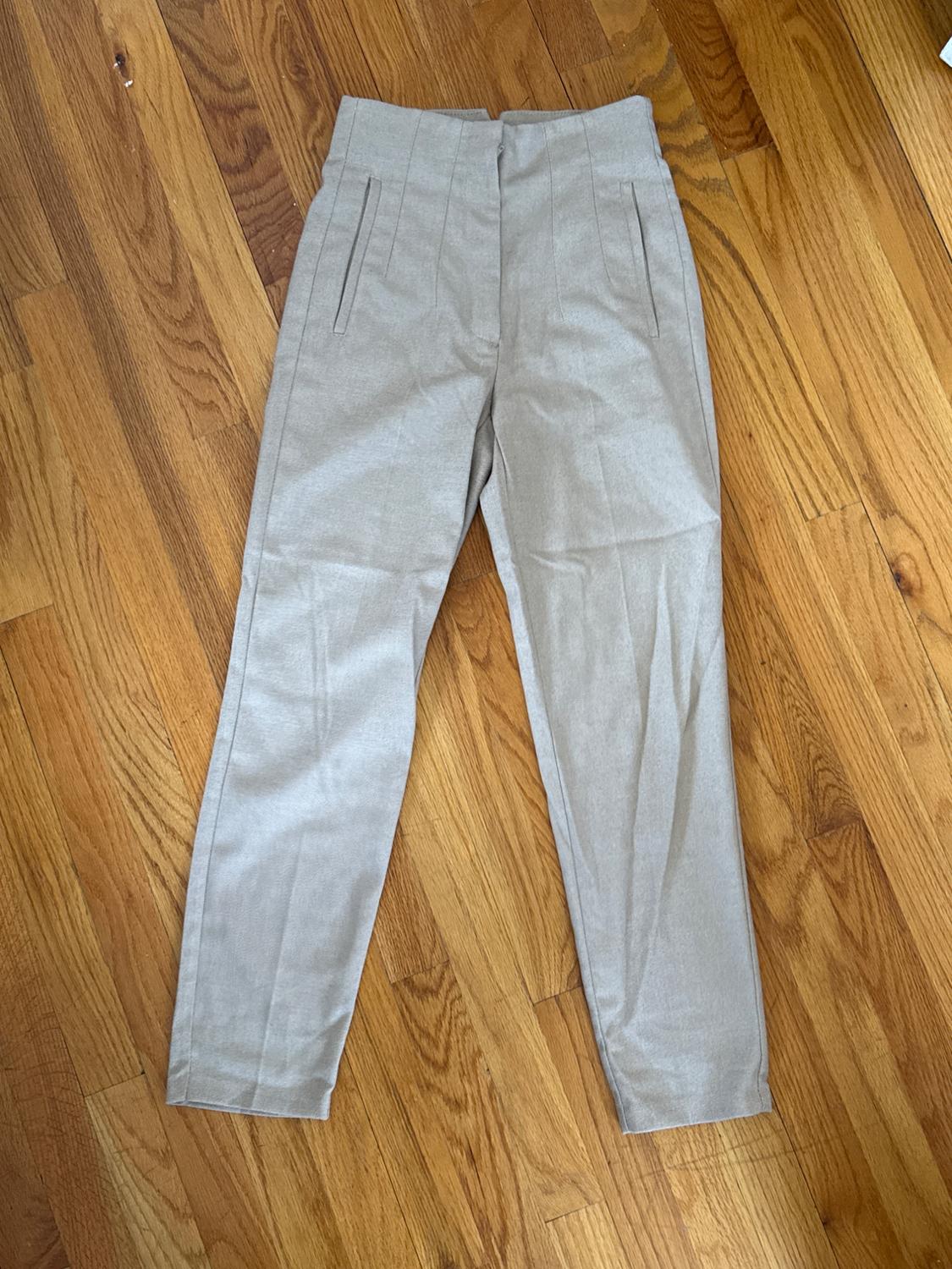 Light beige fabric pants with pleats and side pockets, placed on a light wooden floor.