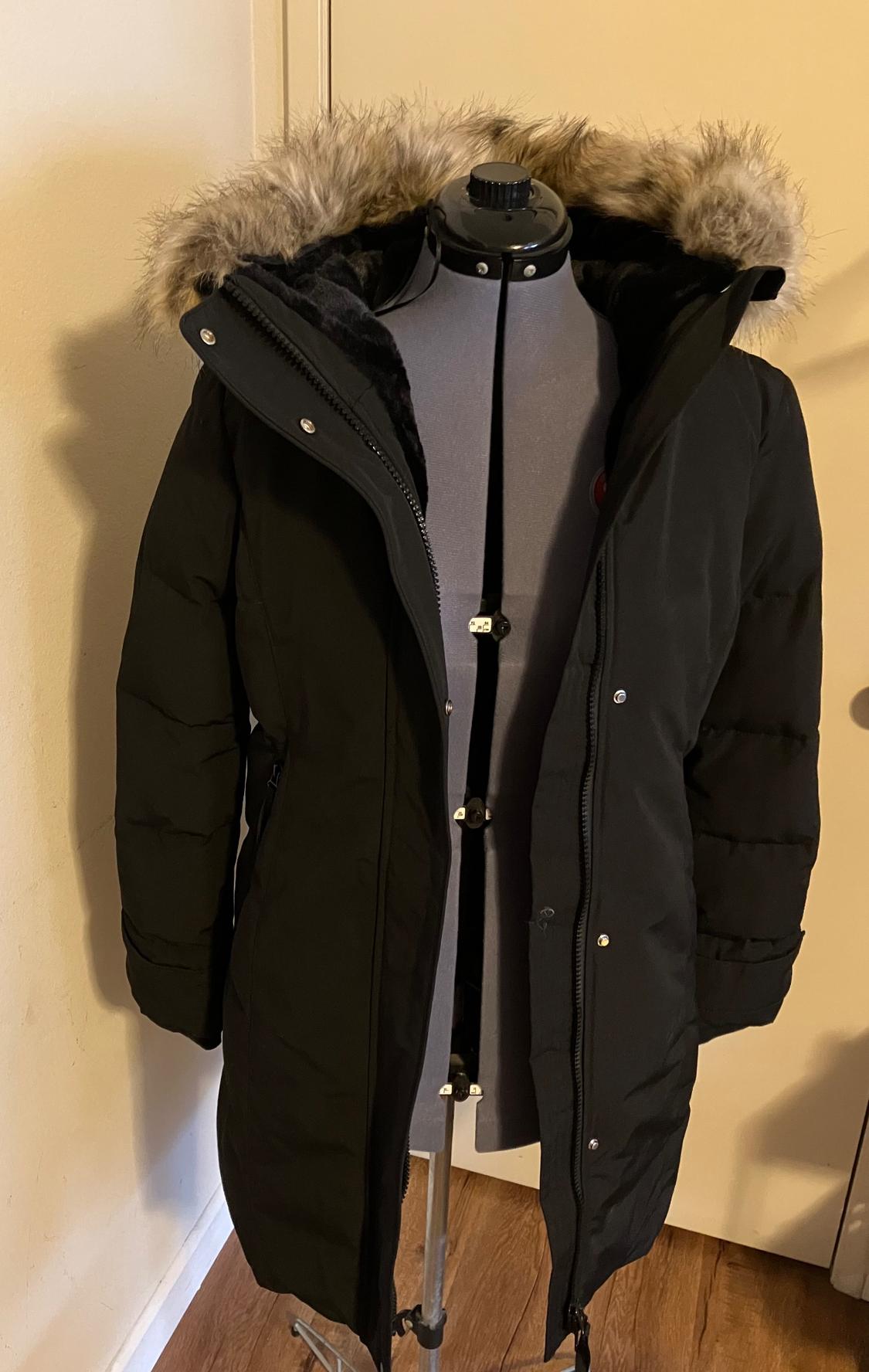 Black coat with faux fur hood on a mannequin, on a wooden floor and beige wall.