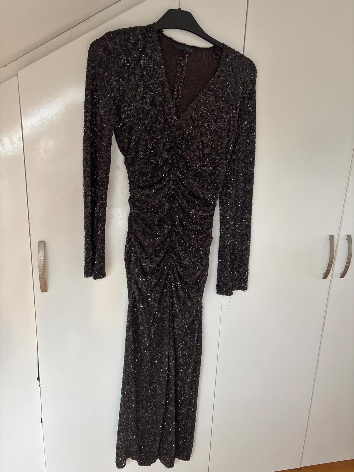 Long black sequinned dress with long sleeves and a V-neck, hanging on a hanger in front of a white wardrobe.