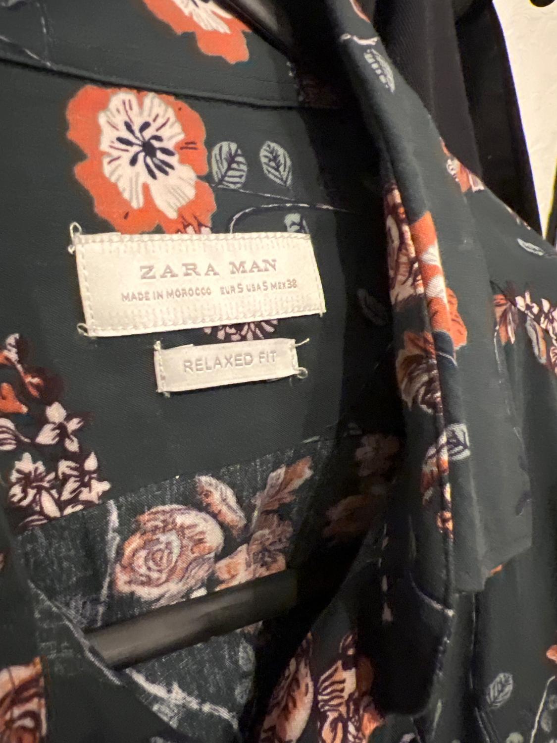 Floral shirt with "ZARA MAN" and "RELAXED FIT" labels, made in Morocco, hanging on a clothes rail. Dark background with orange flowers.