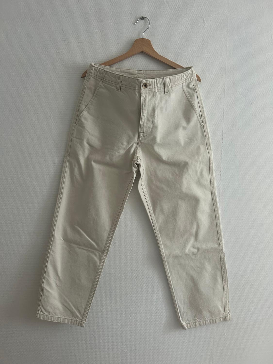 Beige trousers hanging on a wooden hanger against a white wall. Simple design with pockets and a front button.