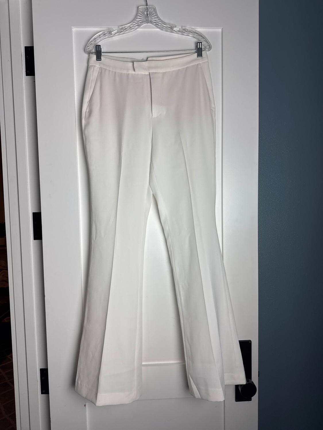 White flared trousers with a front crease, hanging on a transparent hanger over a white door with black hardware.