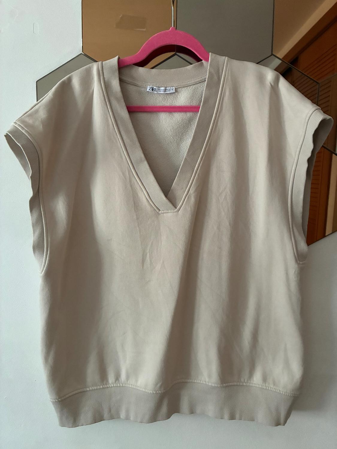 Beige sleeveless V-neck waistcoat, hanging on a pink hanger in front of a hexagonal mirror.