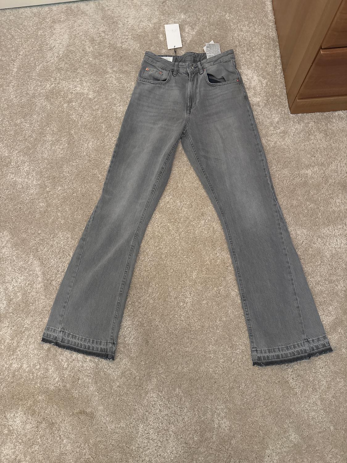 Grey flared jeans on a beige rug, with visible hang tags on the waistband. Wooden chest of drawers to the right.
