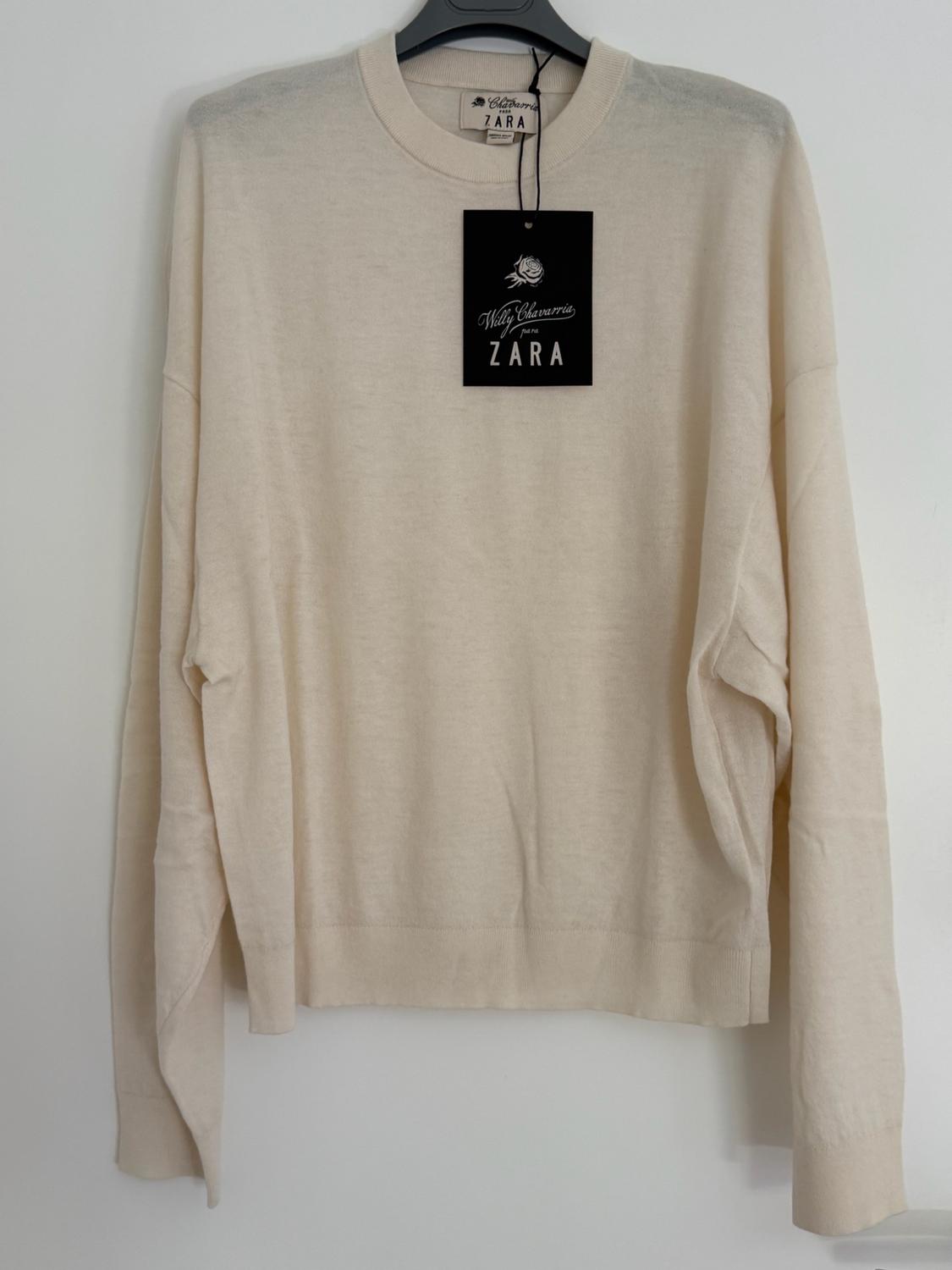 Cream-coloured long-sleeved jumper hanging on a hanger against a white wall. Includes a label bearing the text “Willy Chavarria para ZARA”.