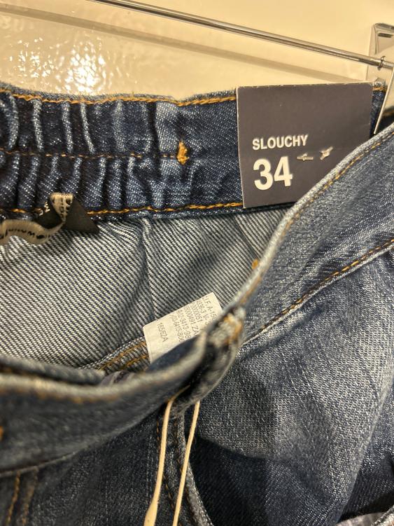 Blue jeans with an elasticated waist and a label that reads "SLOUCHY 34". Stitching detail and visible internal label.