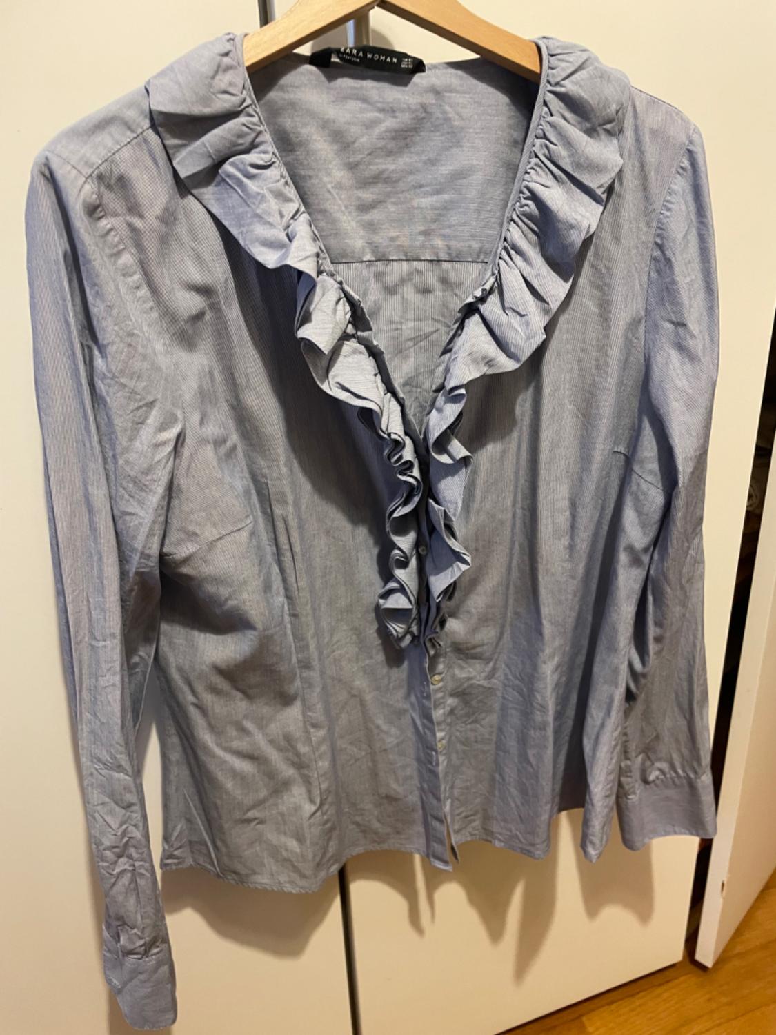 Front view of a blue blouse with frills at the neck and a central button fastening.