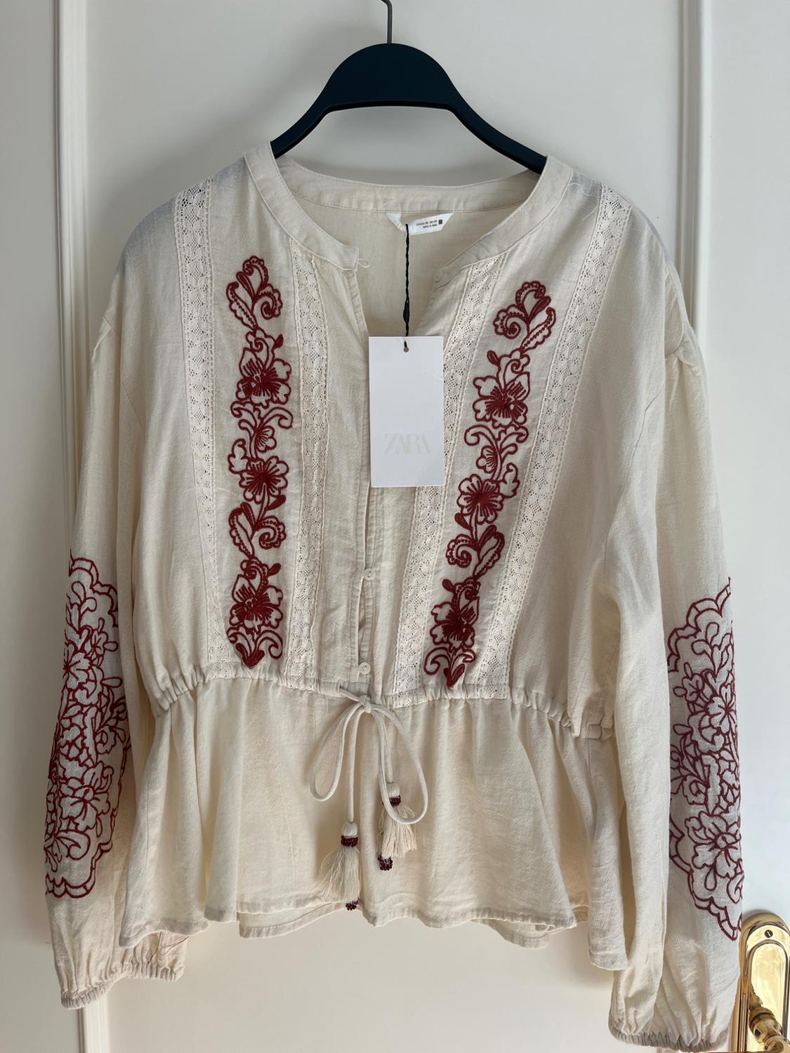 White blouse with red floral embroidery on the front and sleeves, hanging on a black hanger. Visible label with Zara logo.