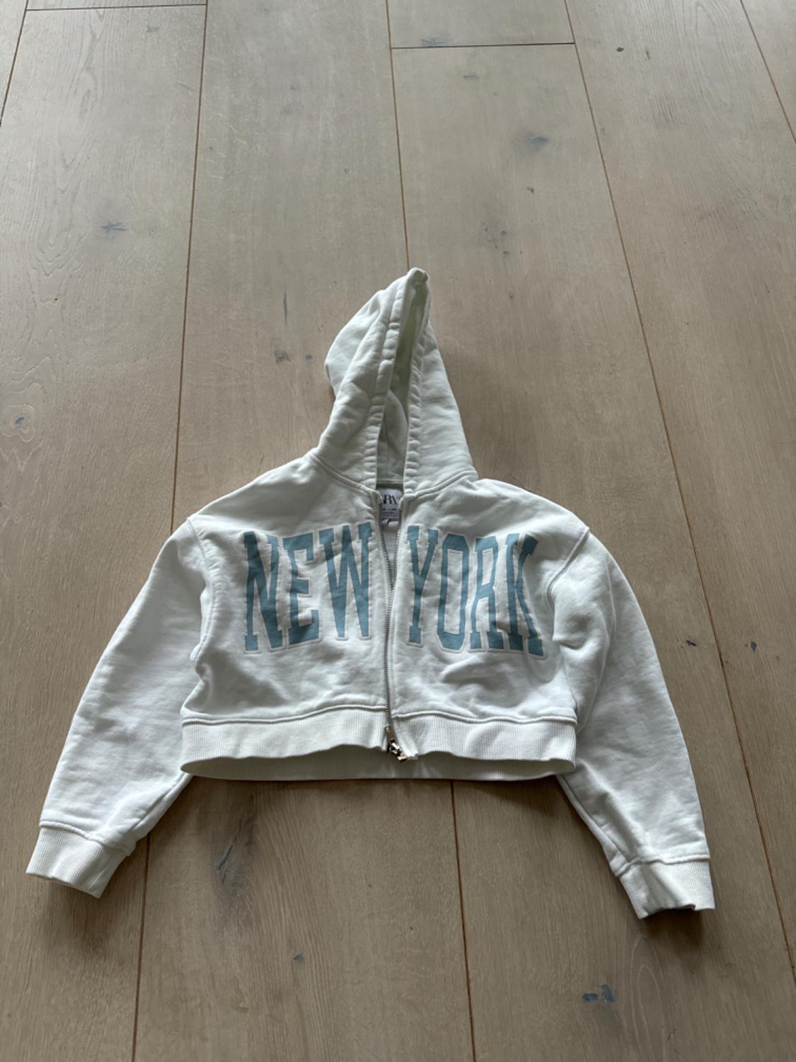 White hooded zip-up sweatshirt, "NEW YORK" text on the front, on a light wooden floor.