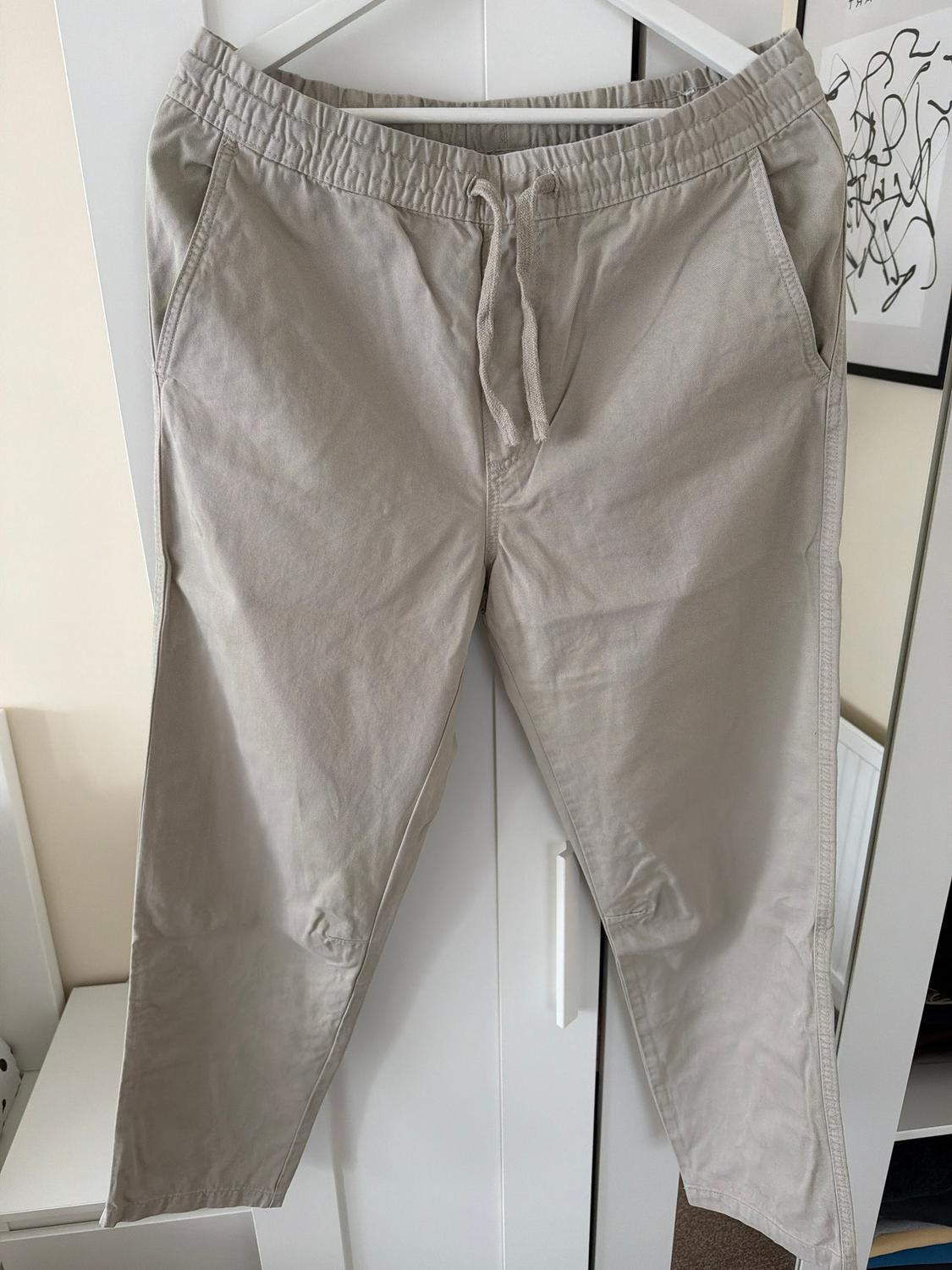 Beige trousers hanging on a white hanger, with an elasticated waist and adjustable drawstring, against a white wardrobe and abstract art background.