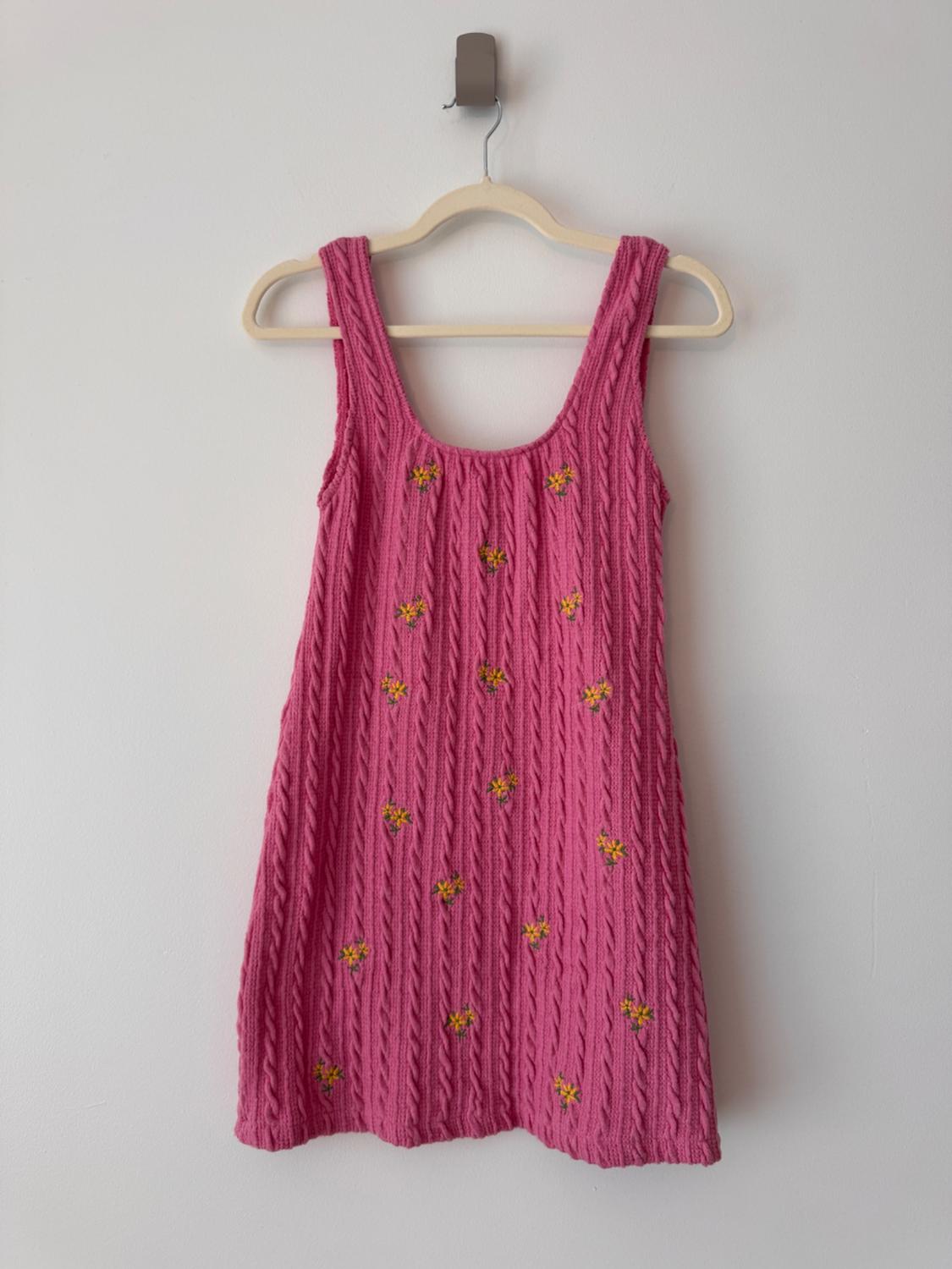 Sleeveless pink knit dress with a braided pattern and small yellow flowers, hanging on a hanger against a white wall.