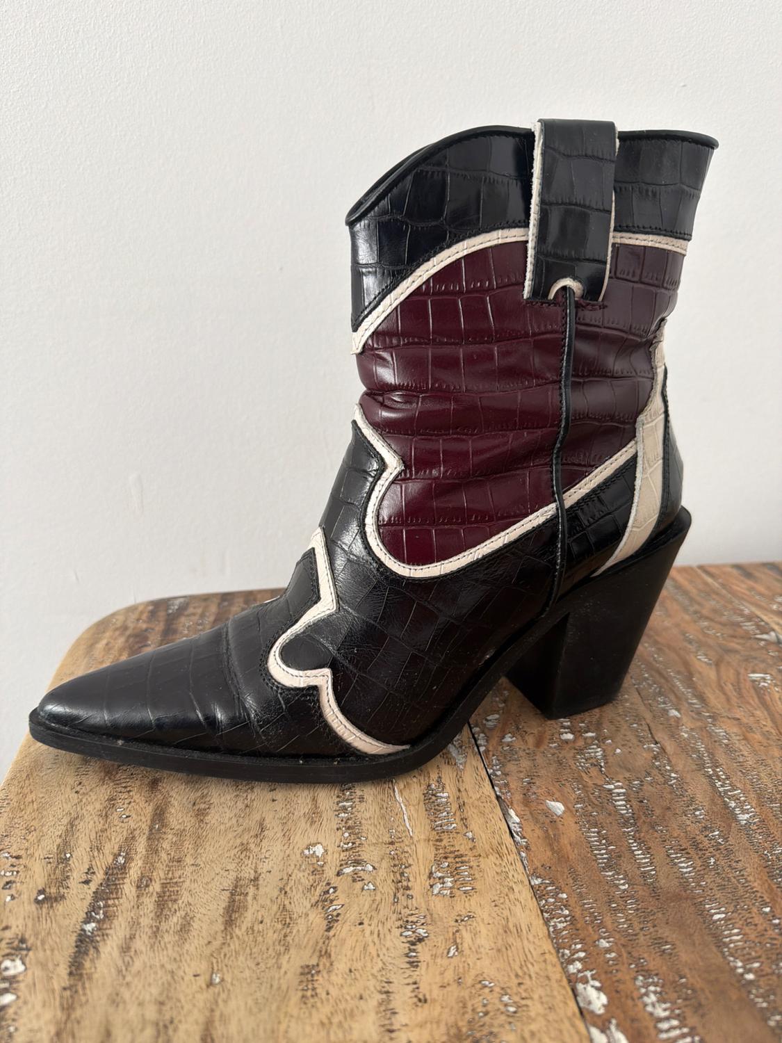 High-heeled boot in black and burgundy textured leather with beige details, on a rustic wooden table.