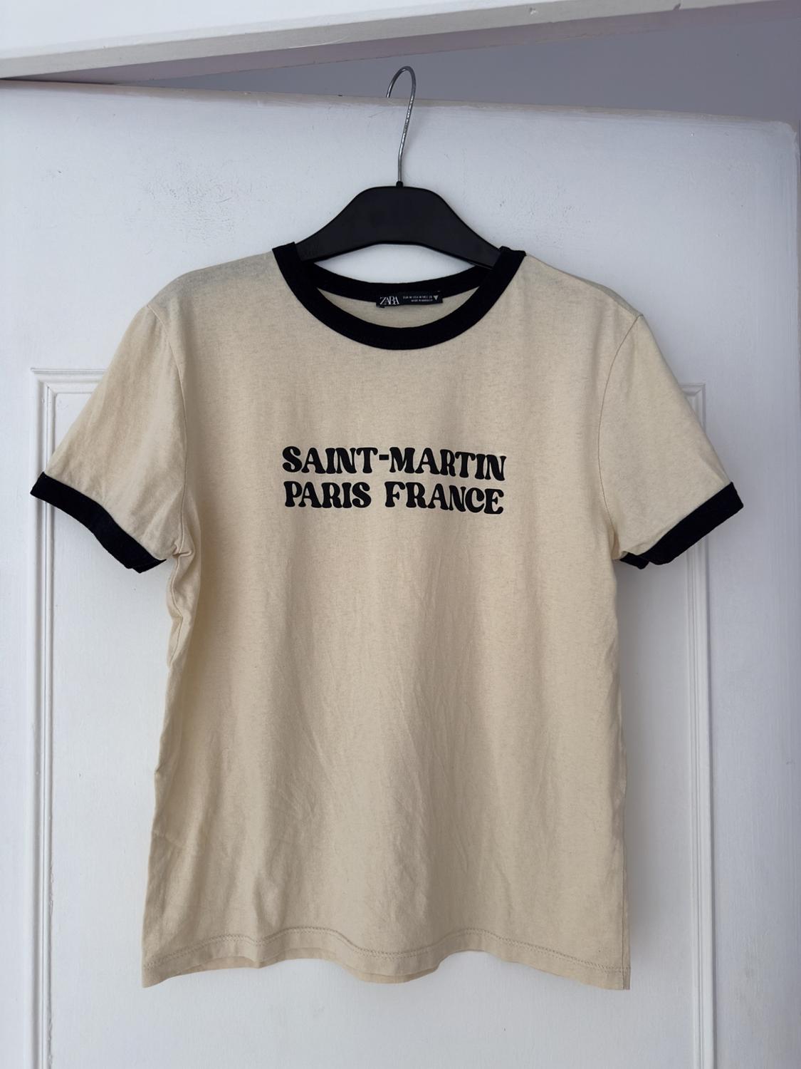 Beige t-shirt with black trim hanging on a hanger. Text on the front: "SAINT-MARTIN PARIS FRANCE". White background.