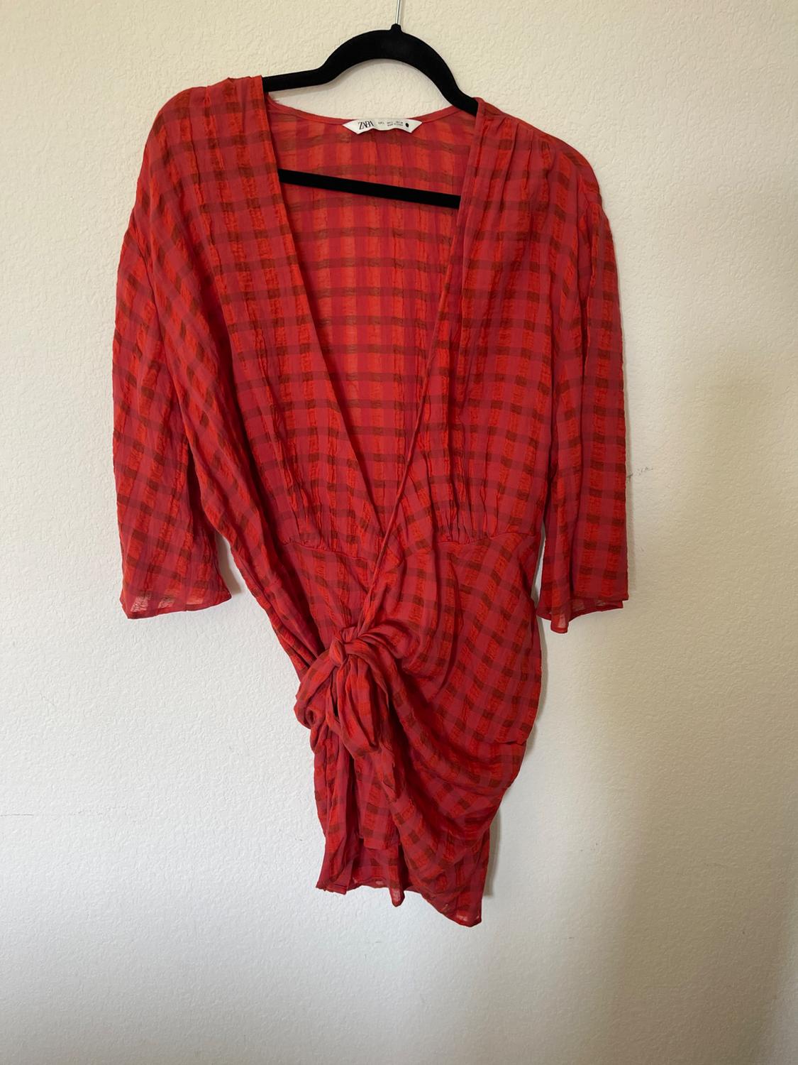 Red dress with a check pattern, three-quarter sleeves and a tie at the waist, hanging on a black coat hanger against a white wall.