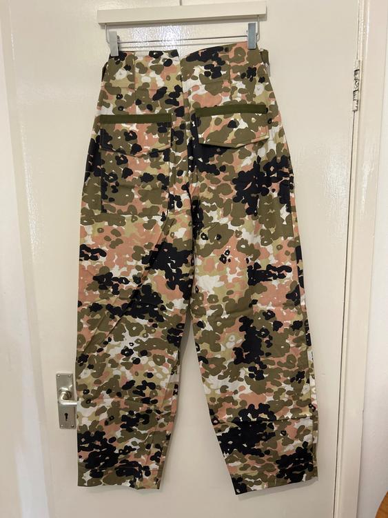 Camouflage print trousers in shades of green, brown, and black, with large back pockets, hanging against a white door background.