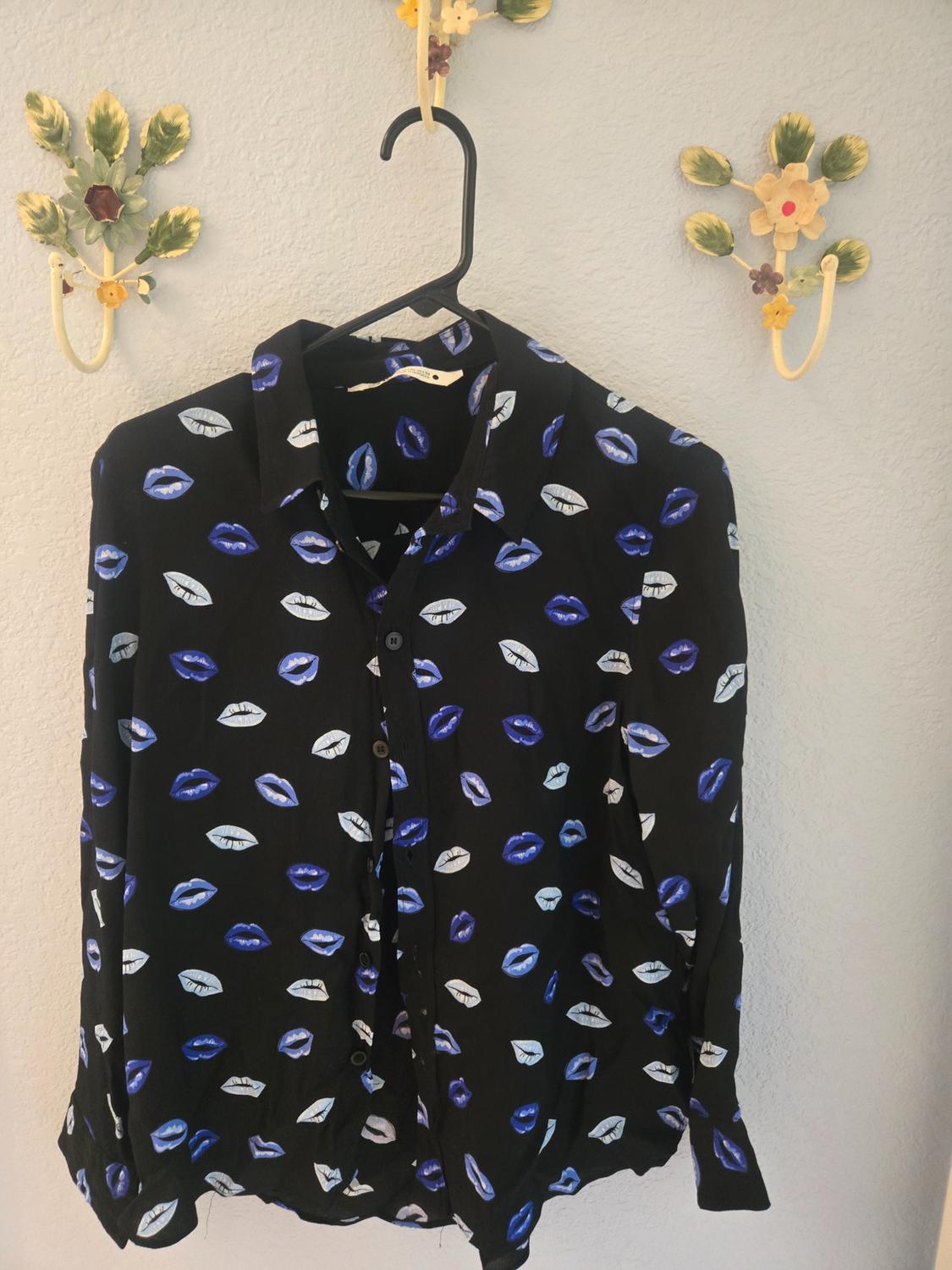 A black shirt with a blue and white lip design is hanging on a black hanger, in front of a wall with decorative flower hooks.