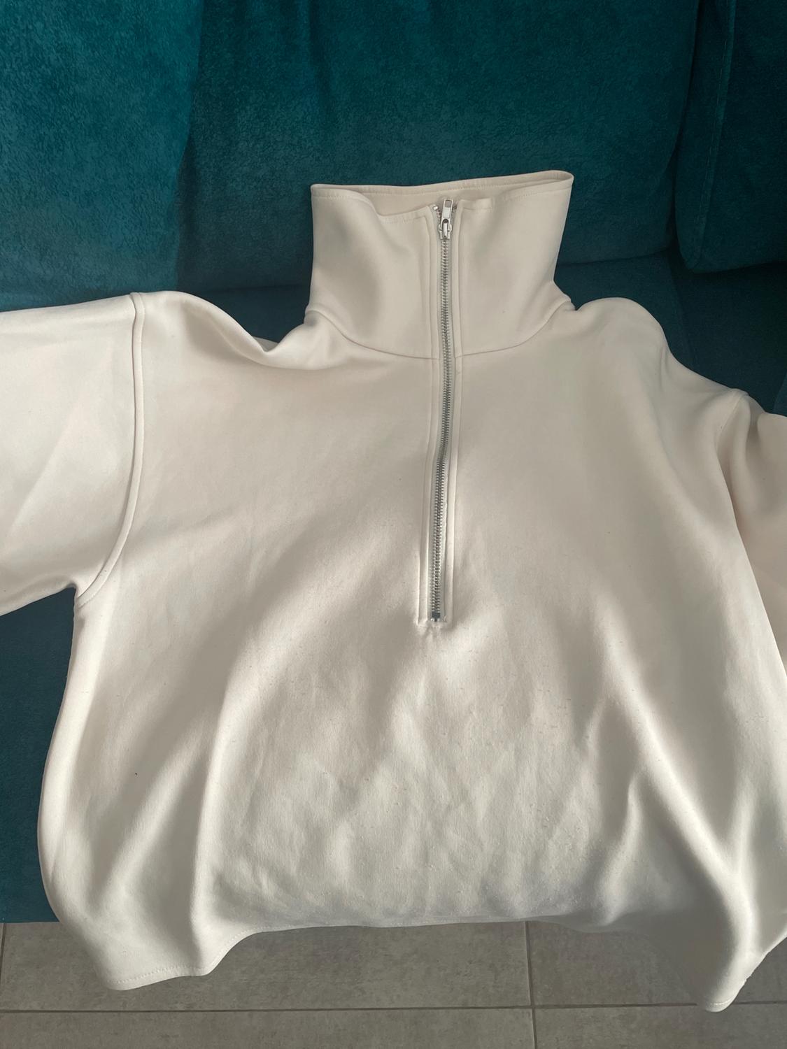 White sweatshirt with a front zip and high neck, placed on a blue sofa. The fabric appears soft and smooth.
