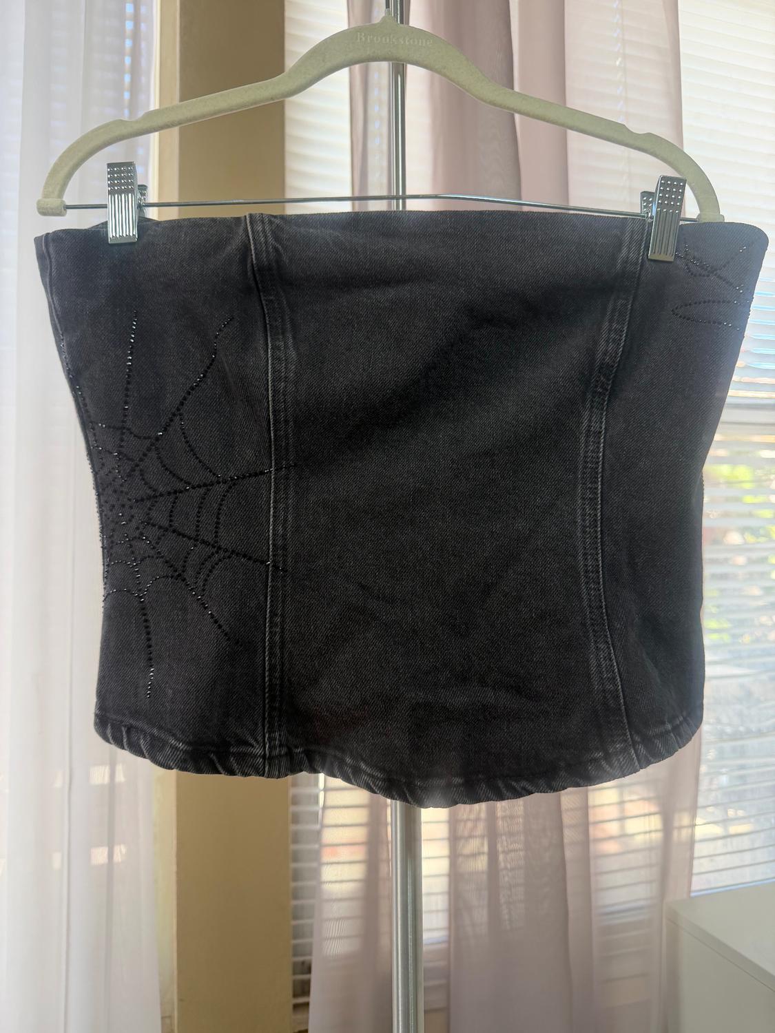 Black denim top with a sequined spiderweb design, hanging on a Brookstone hanger in front of light curtains.