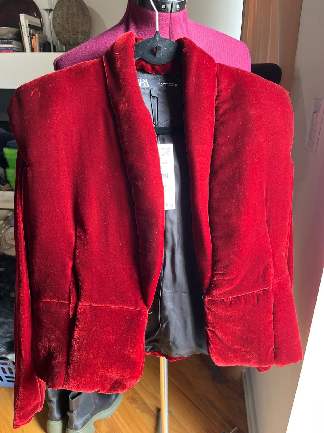 Red velvet jacket on a purple mannequin, with visible tag. Background with shelving and natural light entering from the right.