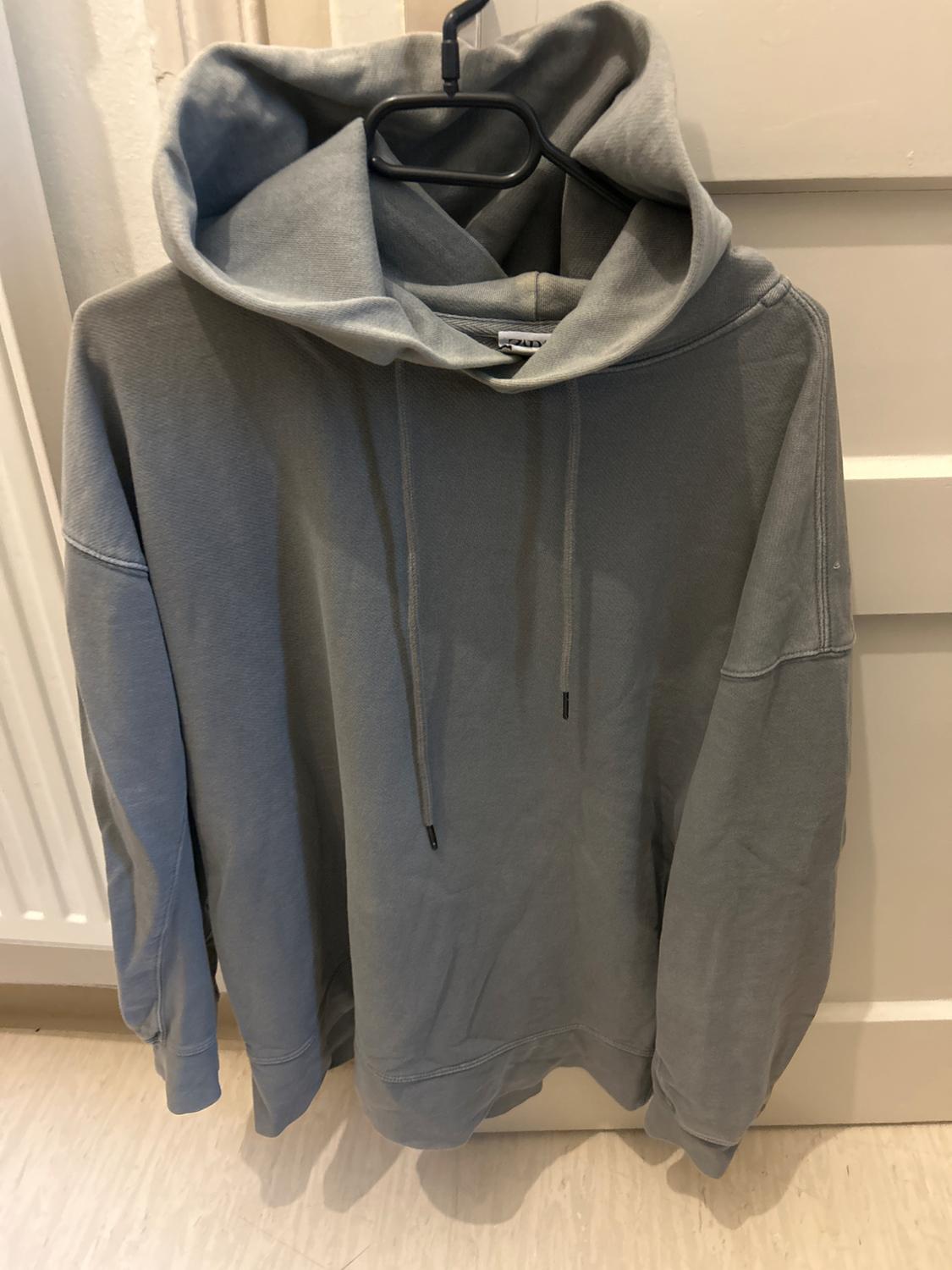 Grey hooded sweatshirt with drawstrings, hanging on a black hanger, against a white door and light-coloured wall background.