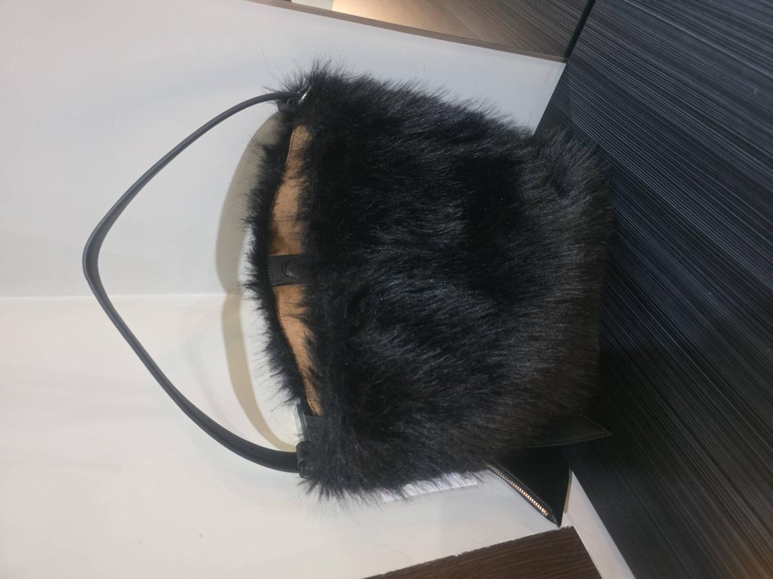 Black faux fur handbag with a thin strap on a white shelf, next to a dark textured panel. Its brown interior is visible.