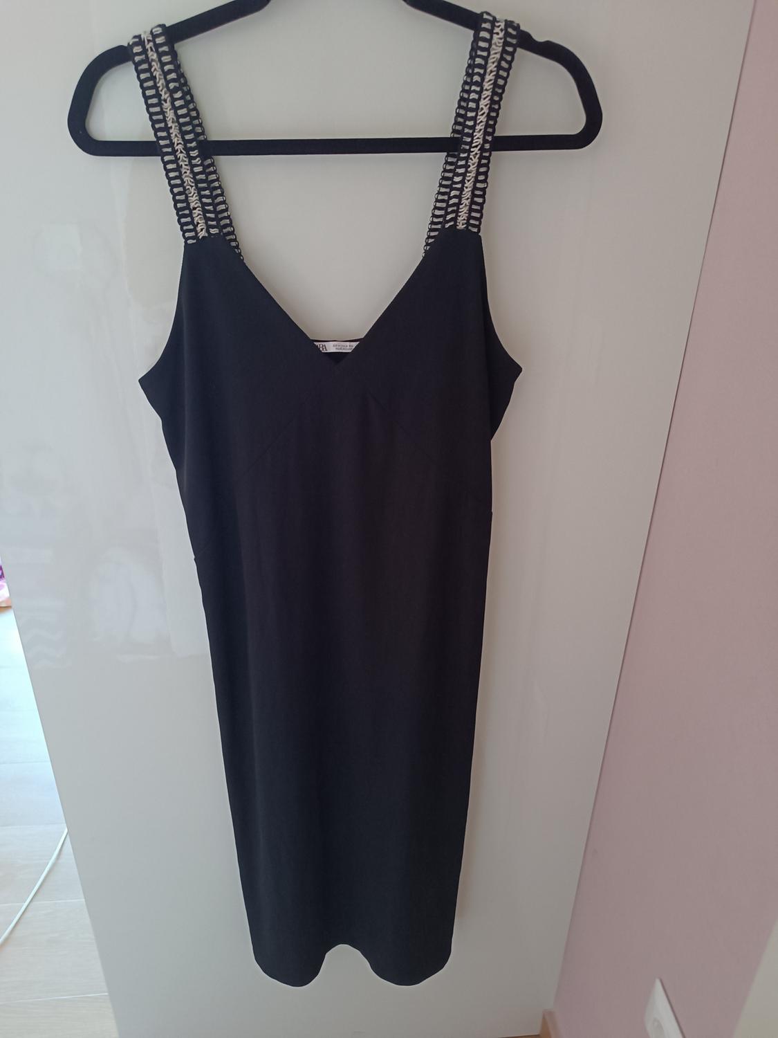 Black sleeveless dress with straps featuring a black and white interwoven design, hung on a black hanger over a white door.