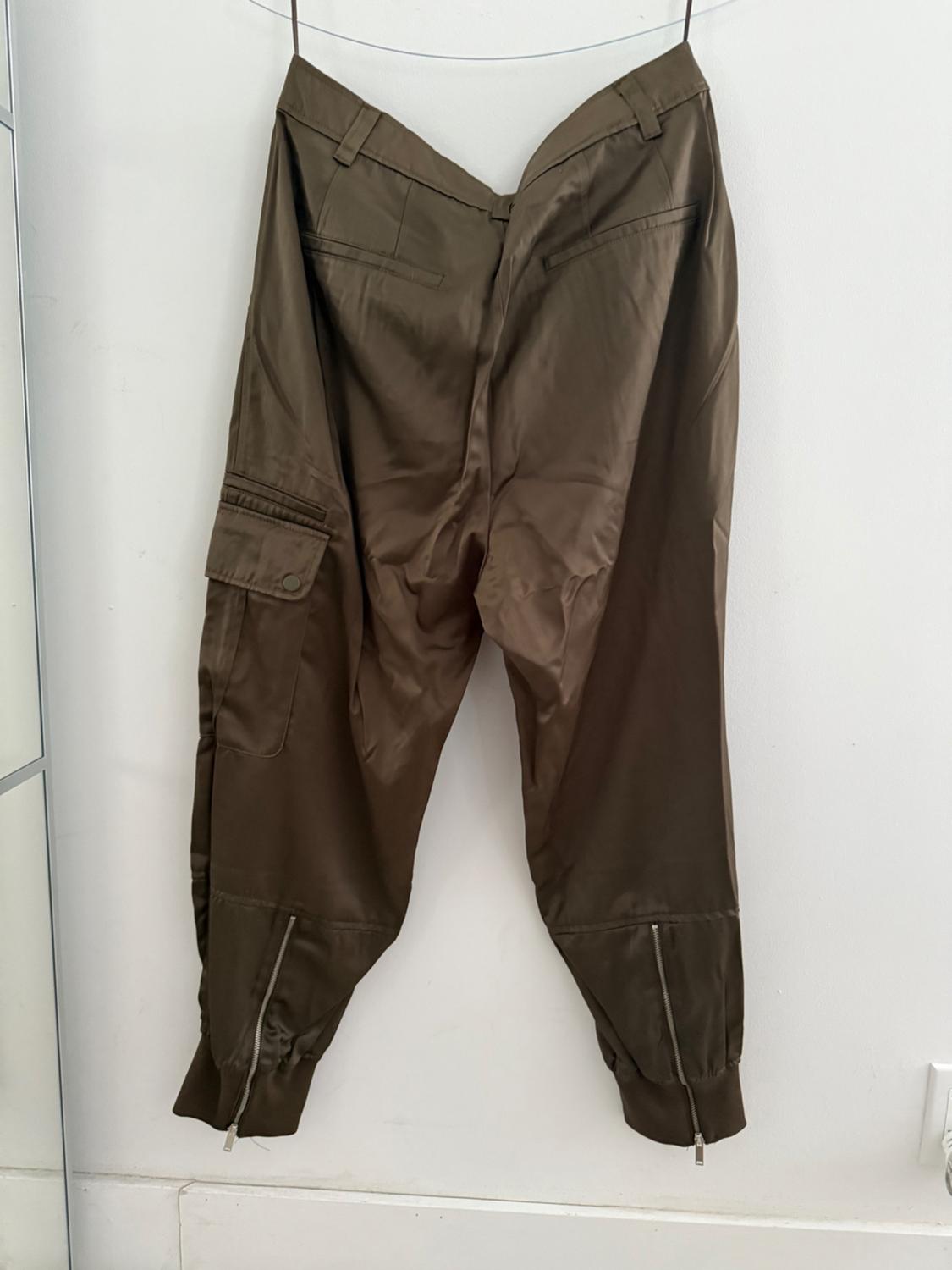 Olive green baggy pants with side pockets and zippers at the ankles, hanging on a thread against a white wall.