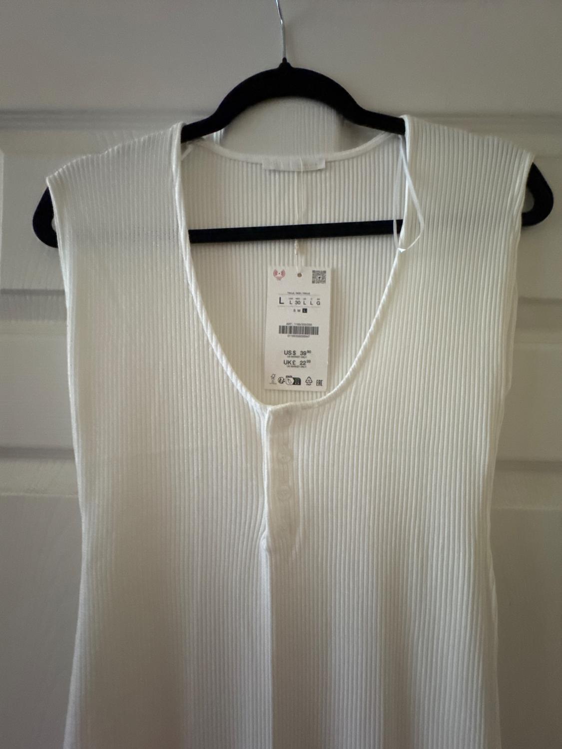 White ribbed t-shirt hanging on a black hanger, with visible tag showing price and size. White door background.
