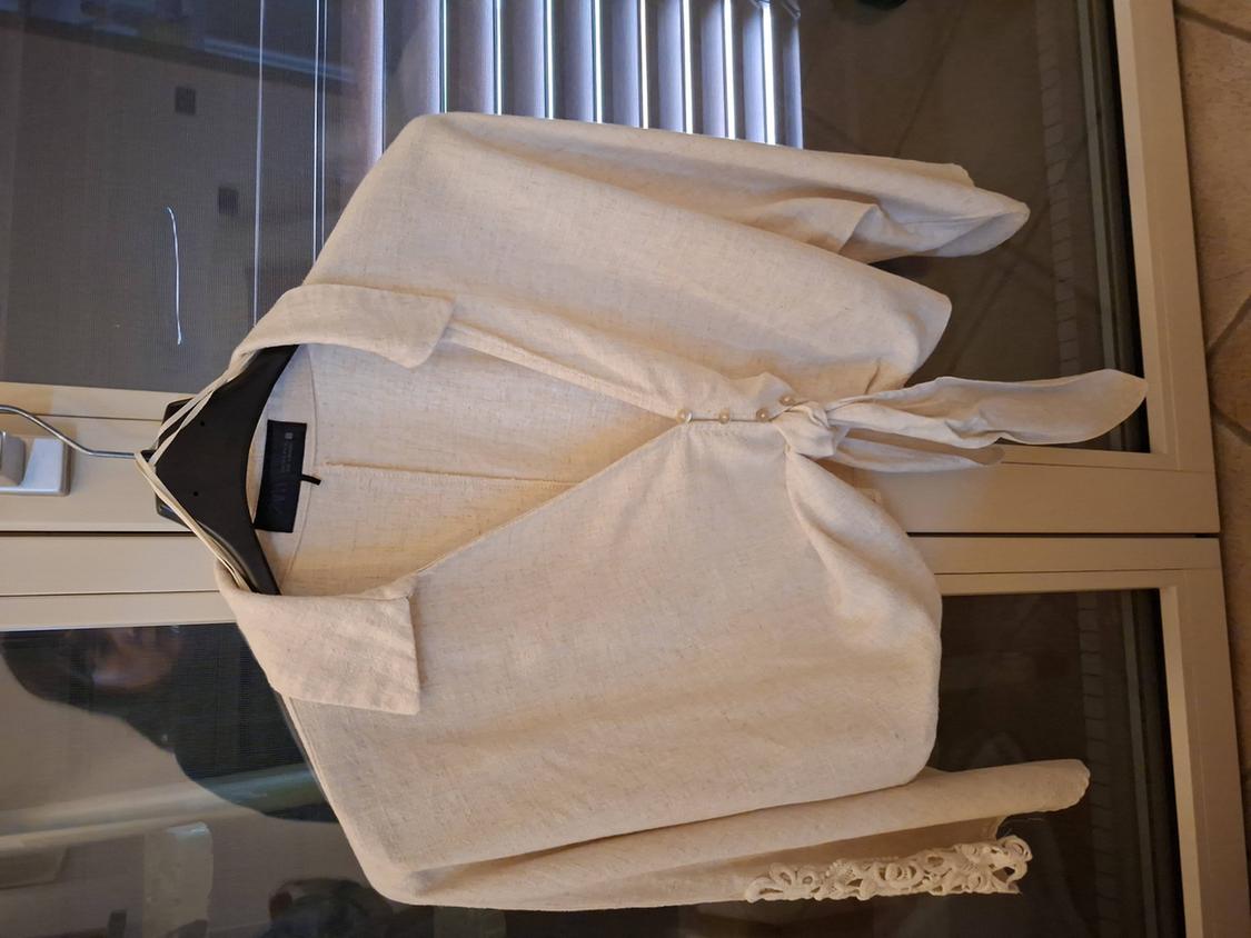 White linen shirt with a front knot, hanging on a black hanger in front of a window with blinds.