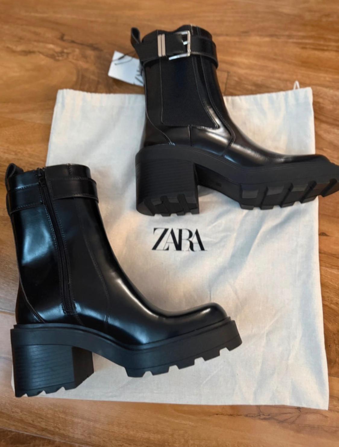 Black patent leather boots with a chunky heel and lug sole on a fabric bag with the "ZARA" logo on a wooden background.