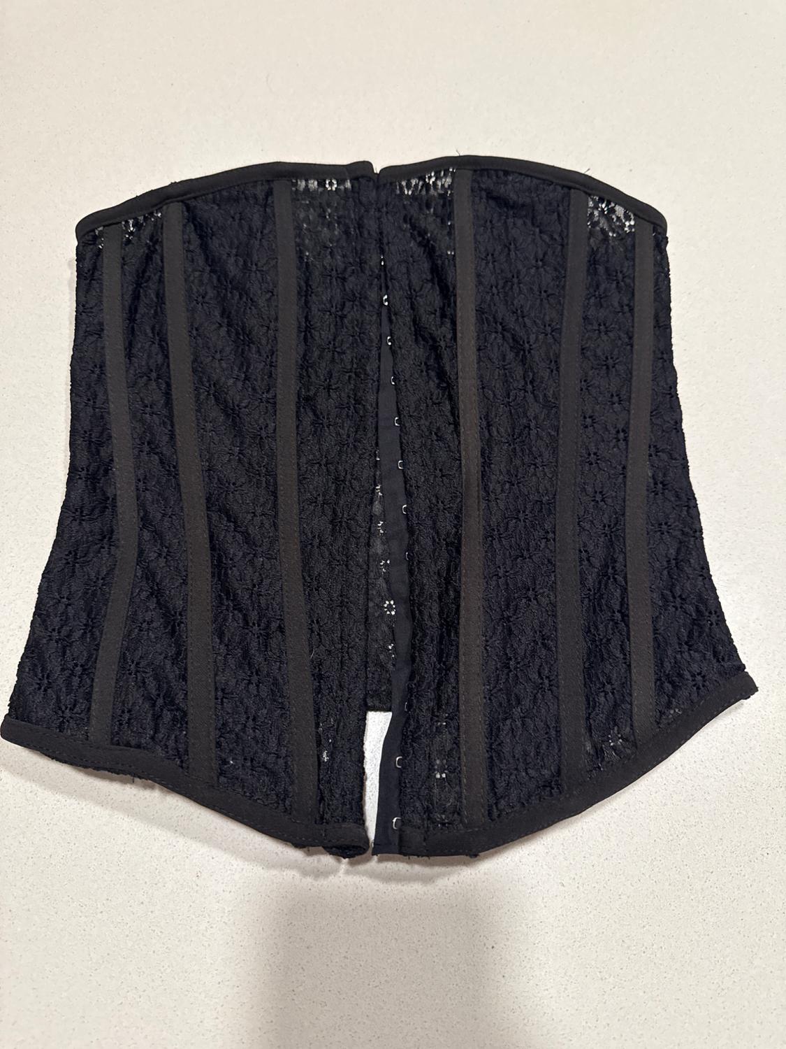 Black lace corset with vertical boning on a light background. Open-front design with hook-and-eye fastening.