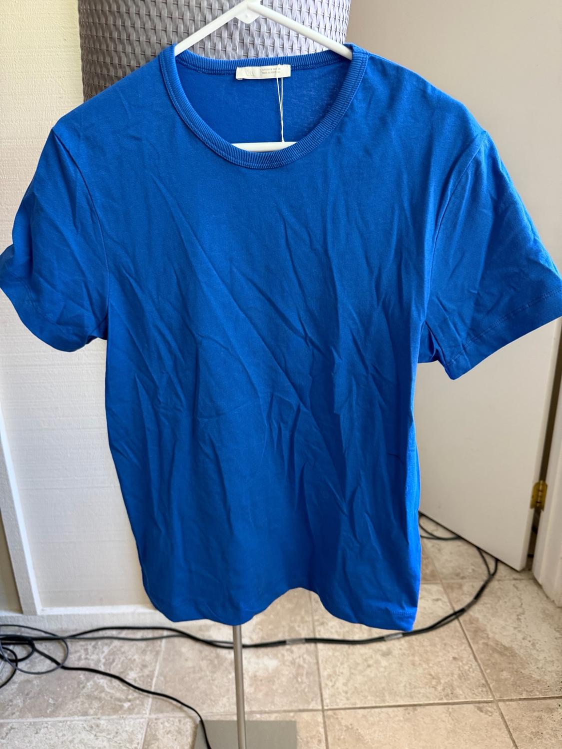 Short-sleeved blue T-shirt hanging on a white hanger on a metal stand. Indoor background with a tiled floor and cables.
